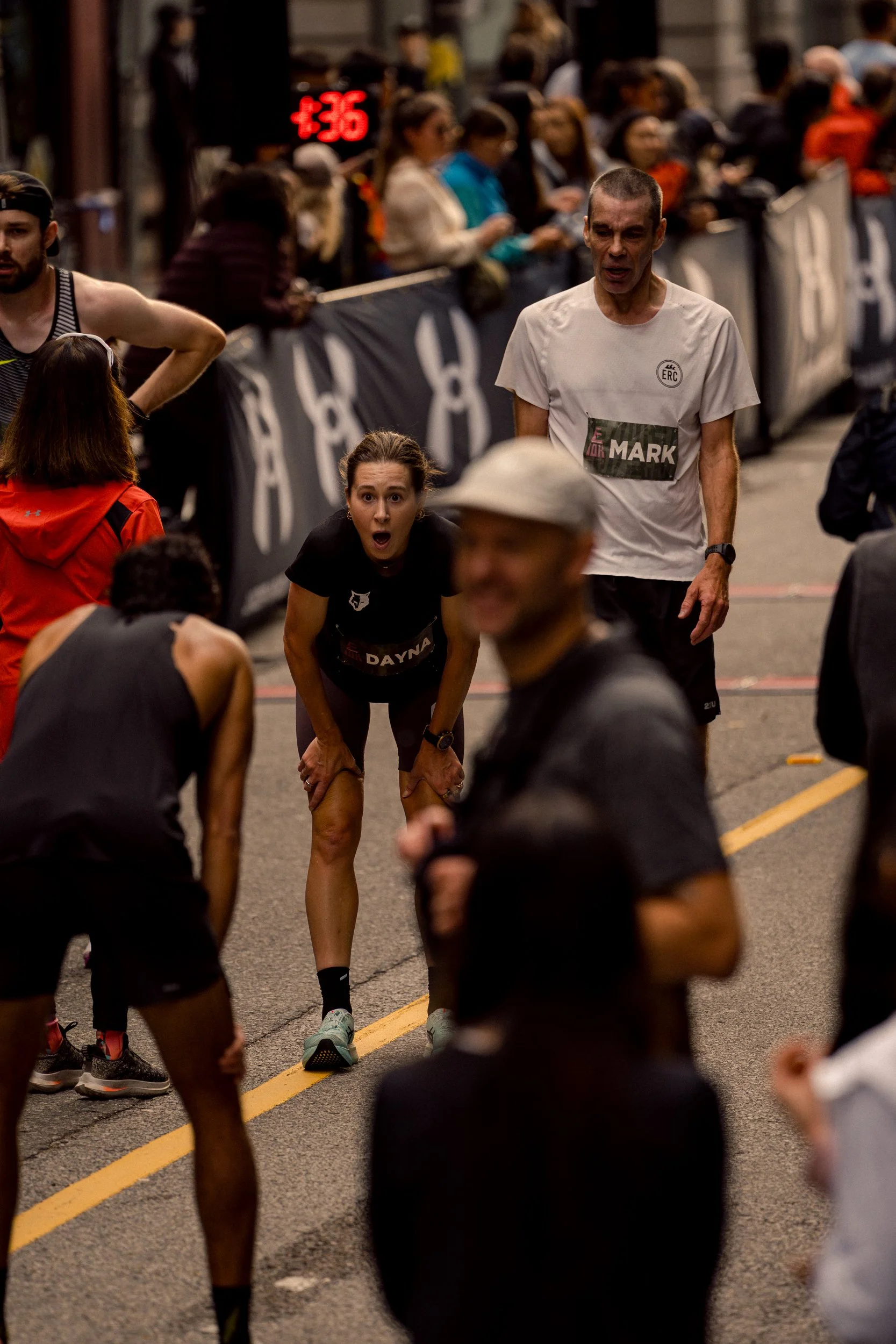 A woman in a black running outfit with a surprised or tired expression during a race, leaning forward while other runners and spectators are present in the background.