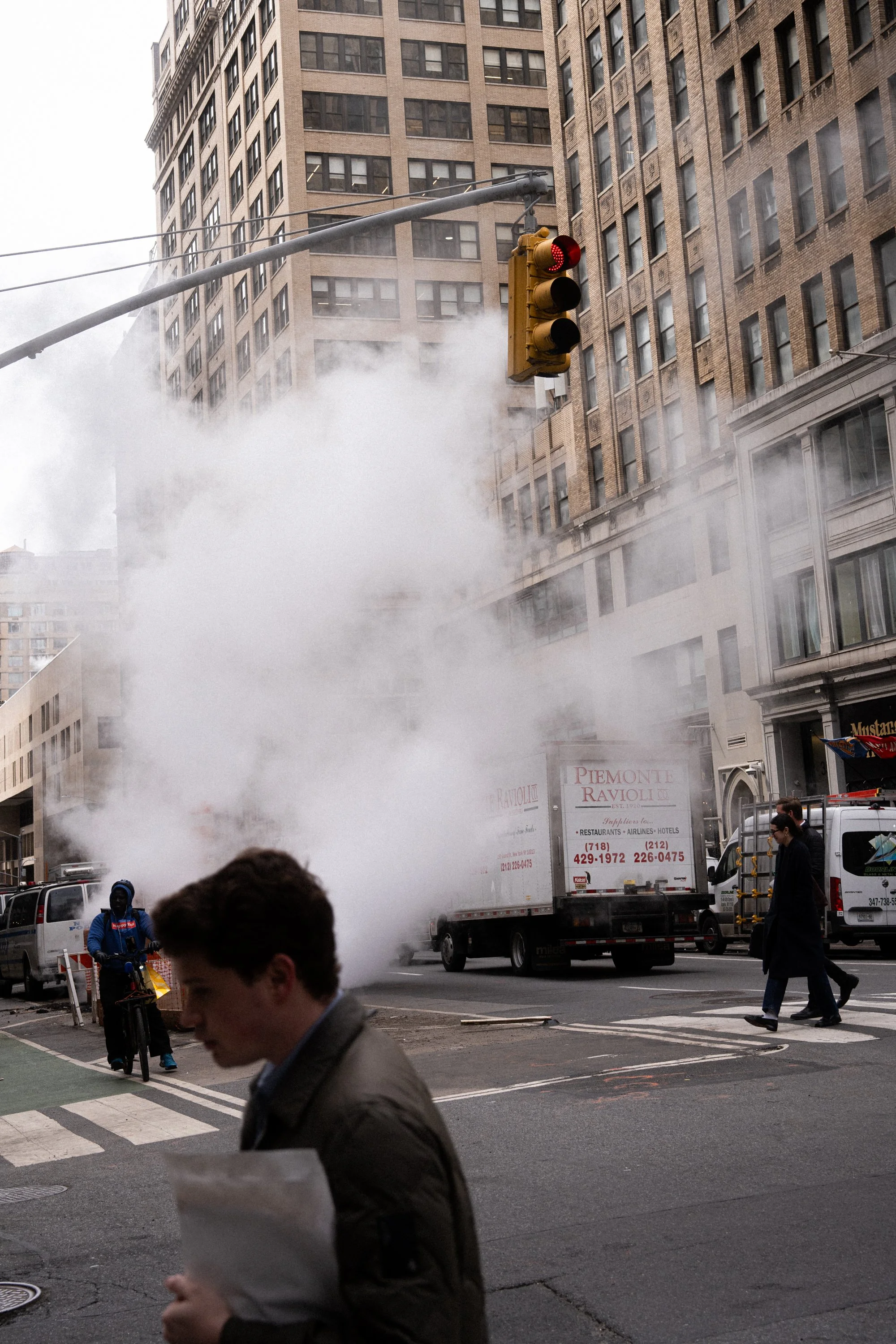 City street scene with a red traffic light, steam rising from a manhole, people walking, and a truck with a sign that reads 'PIEMONTE RAVIOLI' parked on the side of the road with tall buildings in the background.