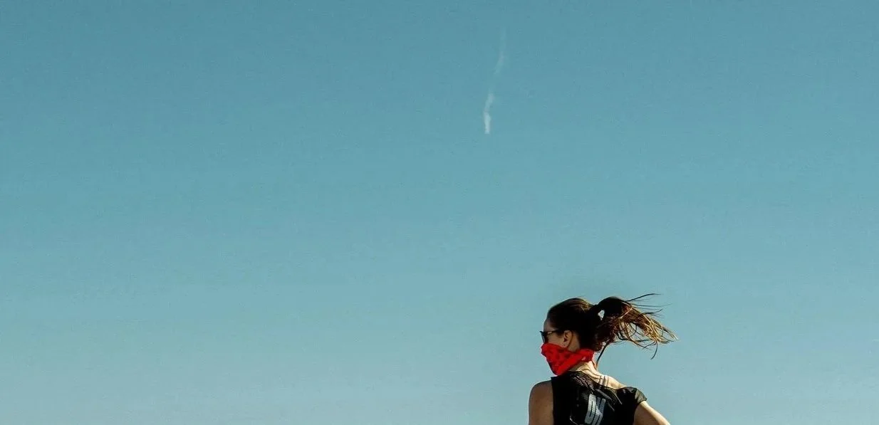 A woman with a ponytail and sunglasses wearing a red face mask, outdoors with a clear blue sky.
