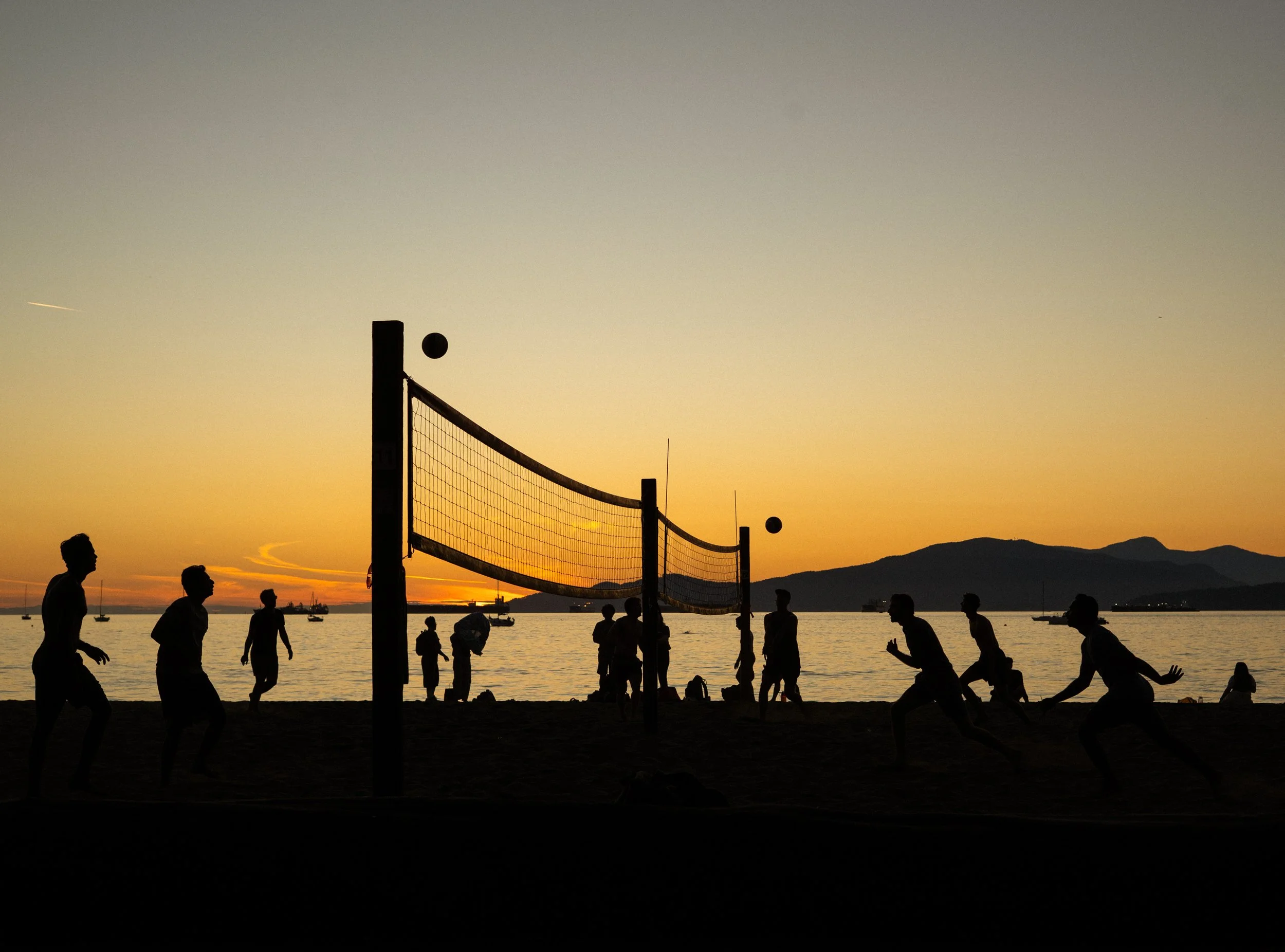 Silhouetted people playing beach volleyball at sunset with mountains in the background and boats on the water.