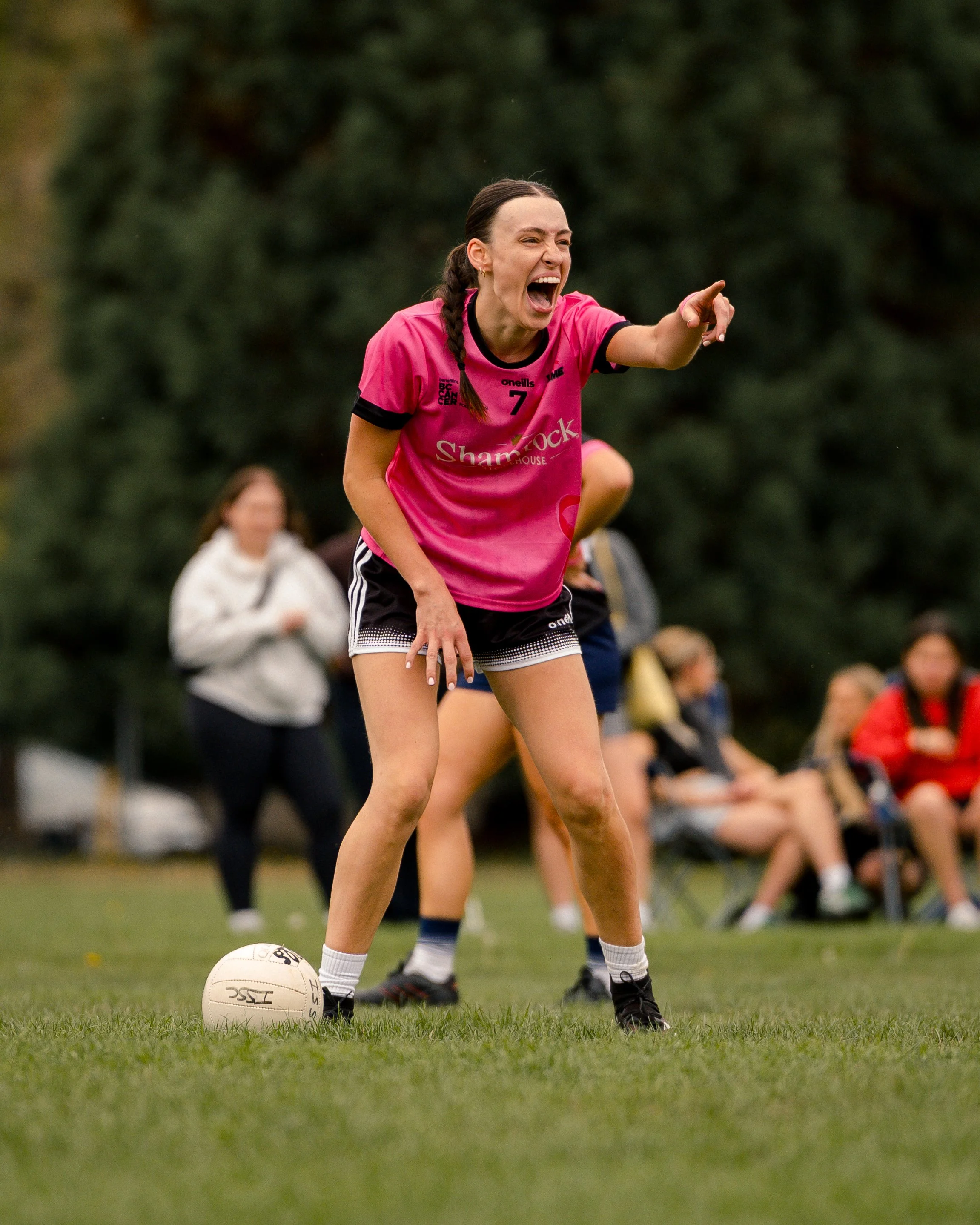 A female soccer player in a pink jersey on the field, pointing and yelling, with spectators in the background.