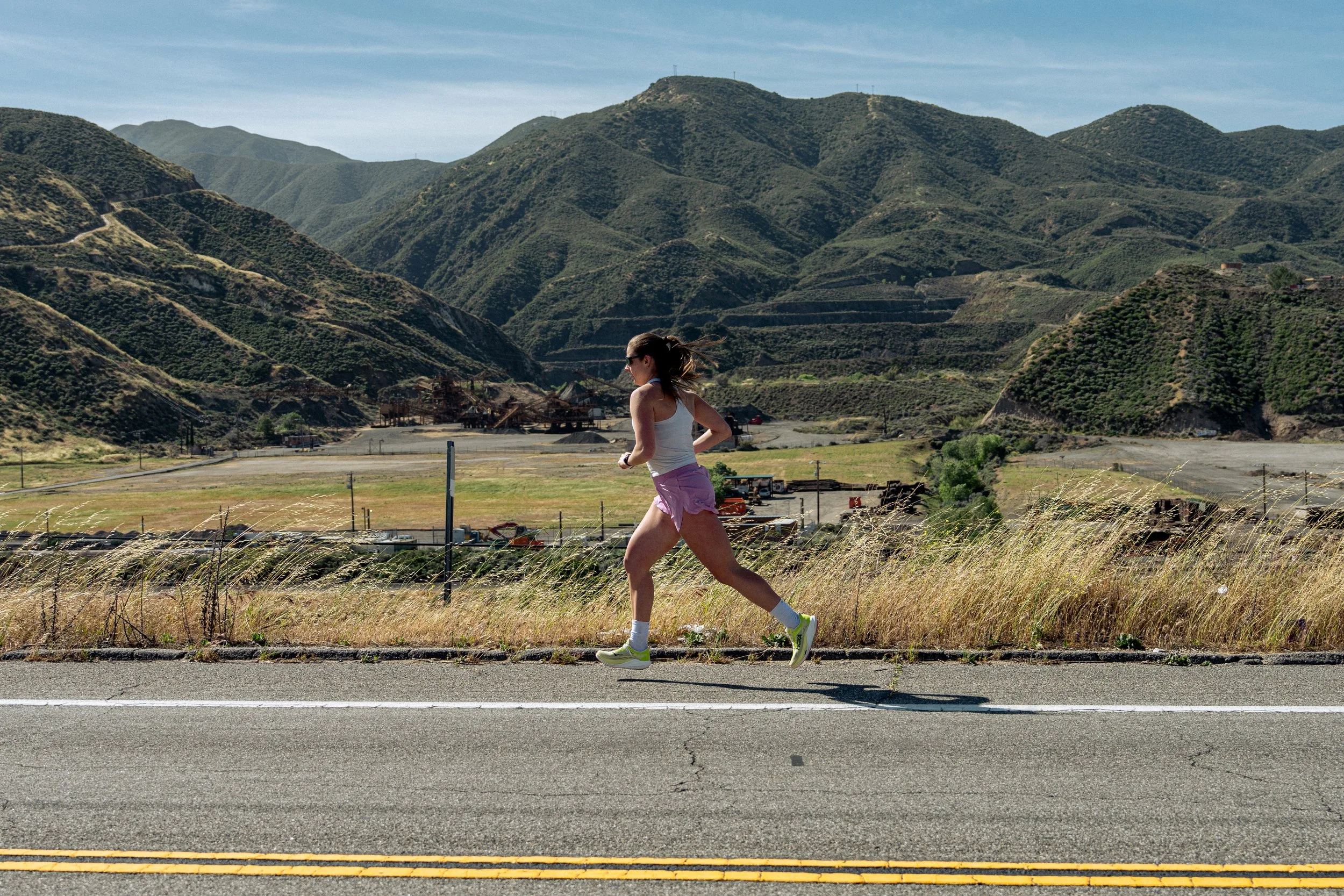 A woman jogging along a roadside with green mountain hills in the background.
