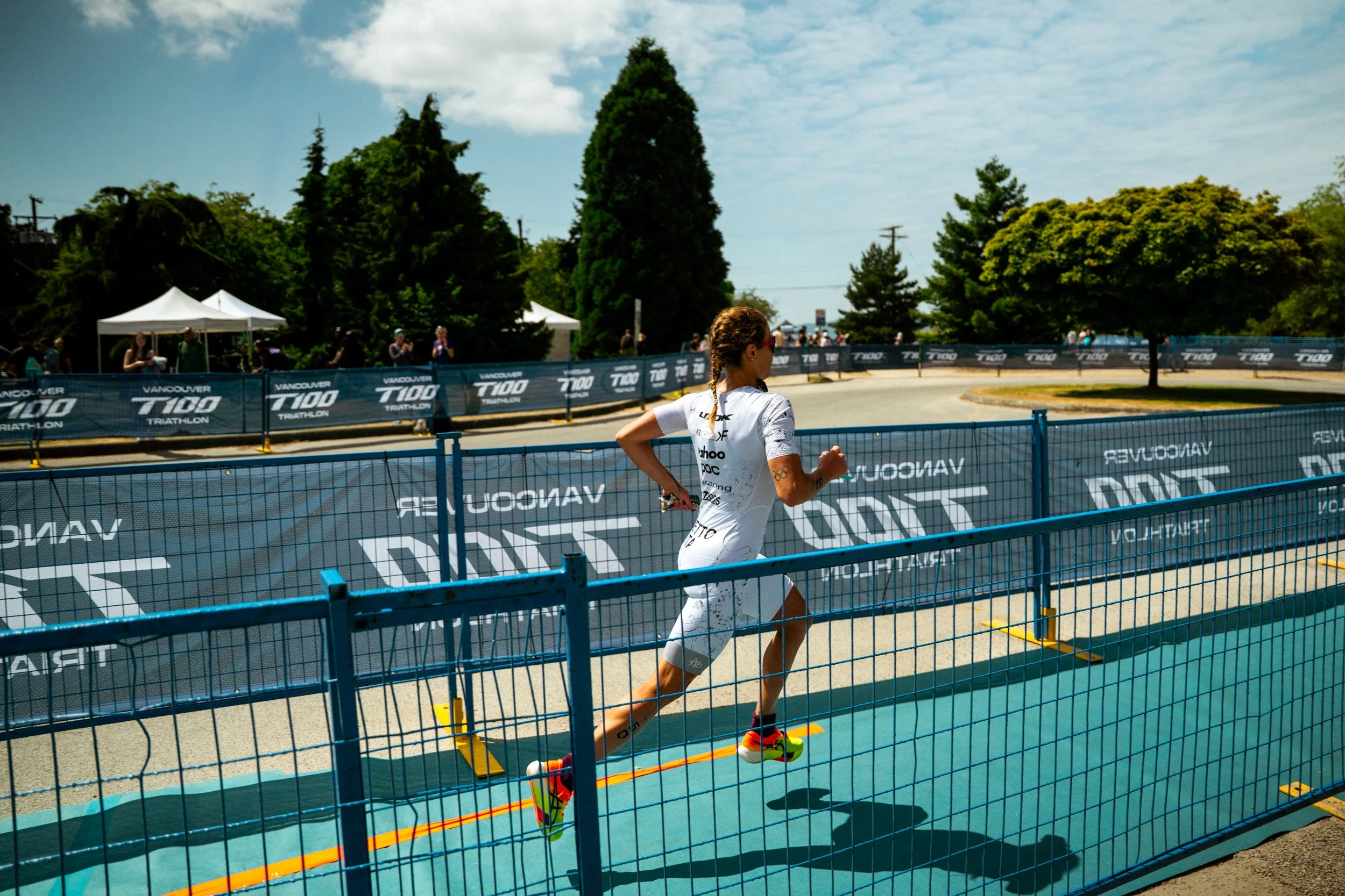 A female athlete running on a track during a triathlon race, wearing a white suit with sponsor logos, sunglasses, and brightly colored running shoes, with a sports armband, alongside a blue safety fence and banners in the background.