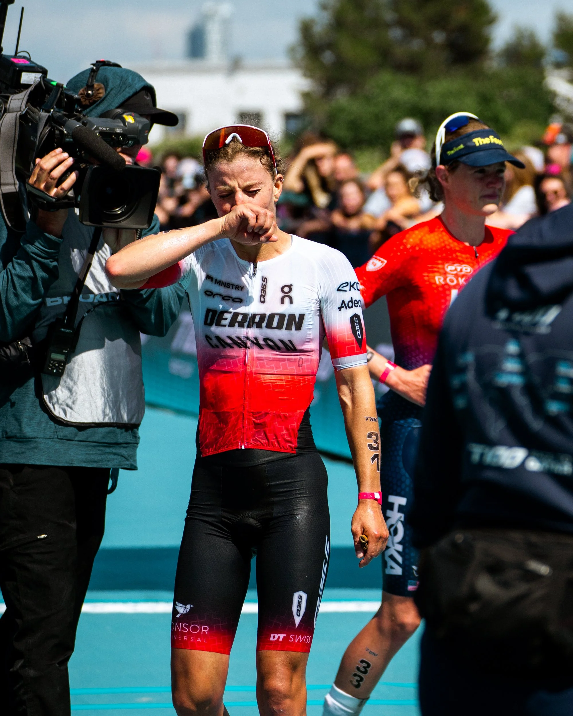 A female cyclist in a red and white Derring Cannon team uniform appears emotional, wiping her face with her hand, after a race. She is surrounded by other athletes, a cameraman, and spectators.
