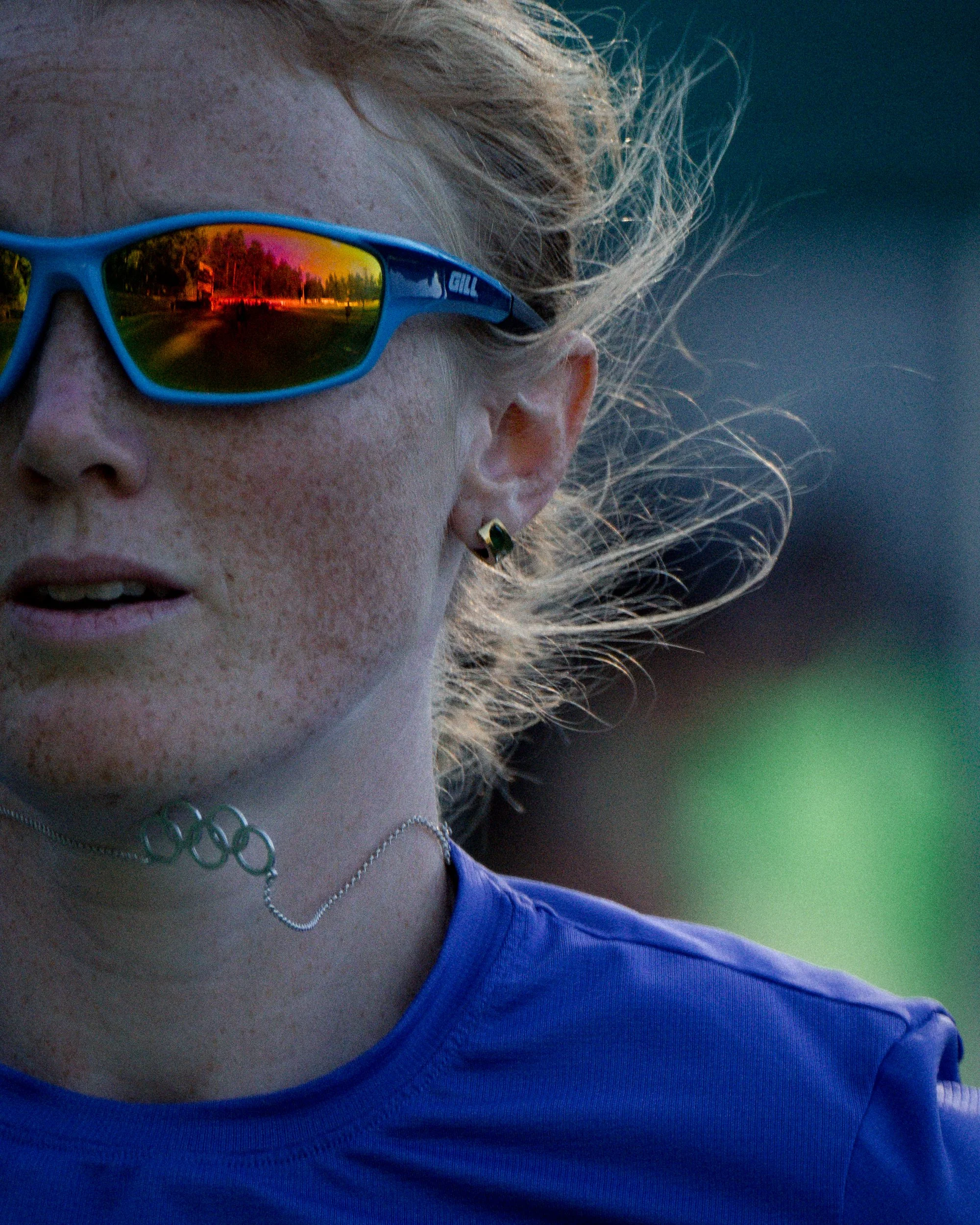 Close-up of a female athlete wearing blue sports sunglasses with reflective lenses, a blue shirt, and Olympic jewelry, outdoors during a sporting event.