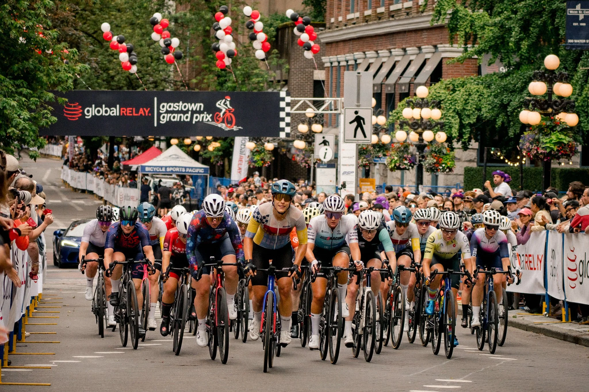 Cyclists starting a race at the Gastown Grand Prix during a street event, with spectators lining the sides, banners, and balloons overhead.