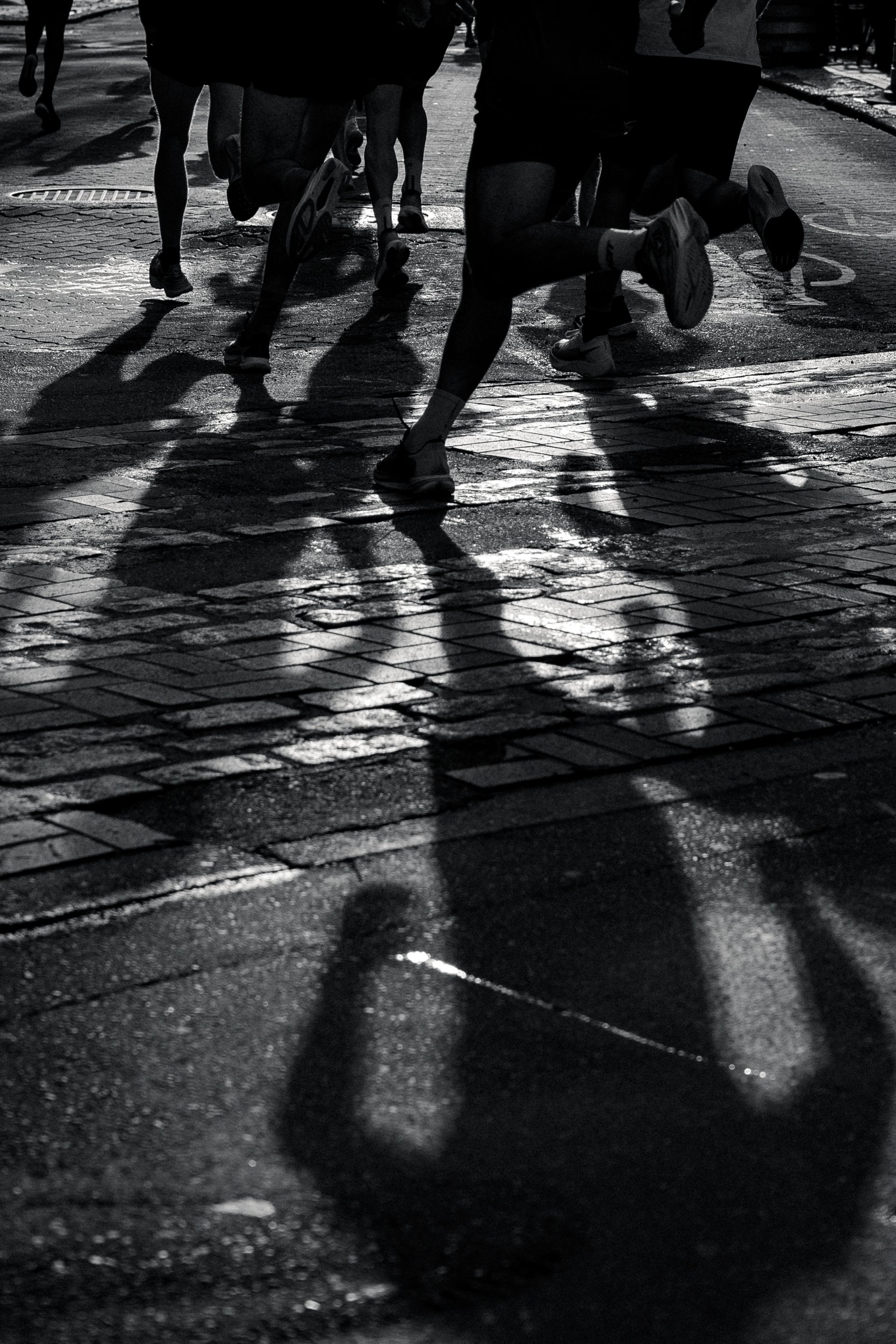 Runners running on a wet city street during daytime, with their shadows cast on the pavement.