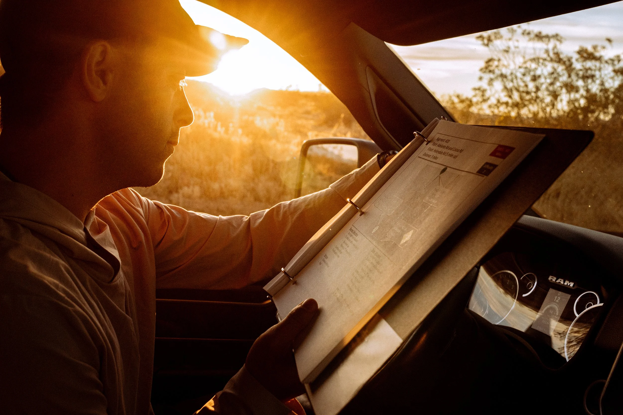 A man in a beige shirt sitting in a car at sunset, reading a clipboard and listening to music through earphones. The sun is setting in the background, creating a warm glow.
