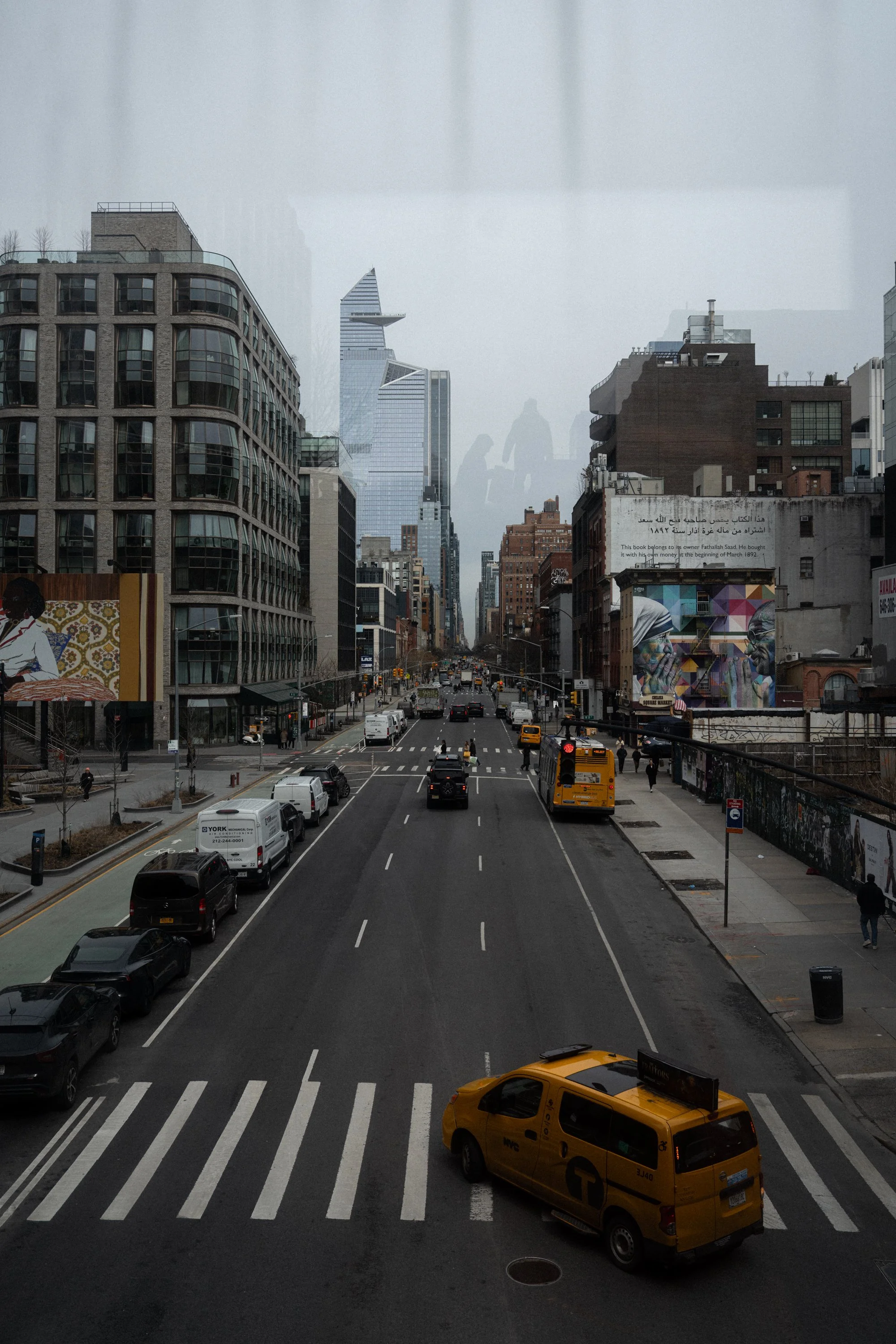 A city street in New York City with traffic, tall buildings, and advertisements. A yellow taxi is in the foreground on a crosswalk, and other vehicles are on the road. Pedestrians are walking on the sidewalks.