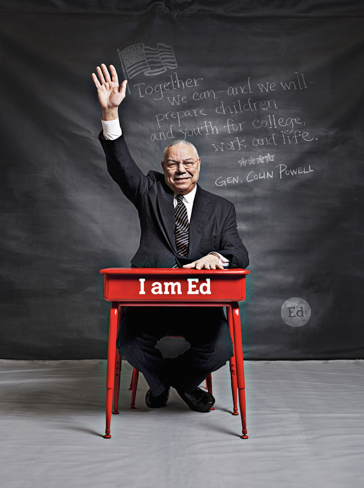 A man in a suit sitting at a red desk with a sign that reads "I am Ed". He is smiling and raising one hand. Behind him on a blackboard is handwritten text about preparing children and youth for college, work, and life, signed by General Colin Powell.