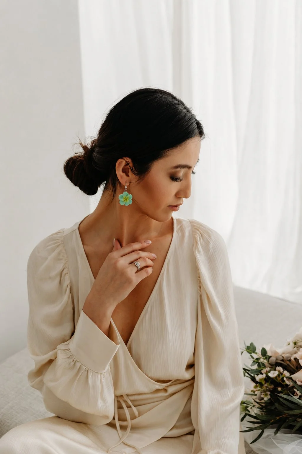 A woman with dark hair in a low bun, wearing a cream-colored dress and flower-shaped earrings, sitting near a window with white curtains, and a bouquet of flowers nearby.