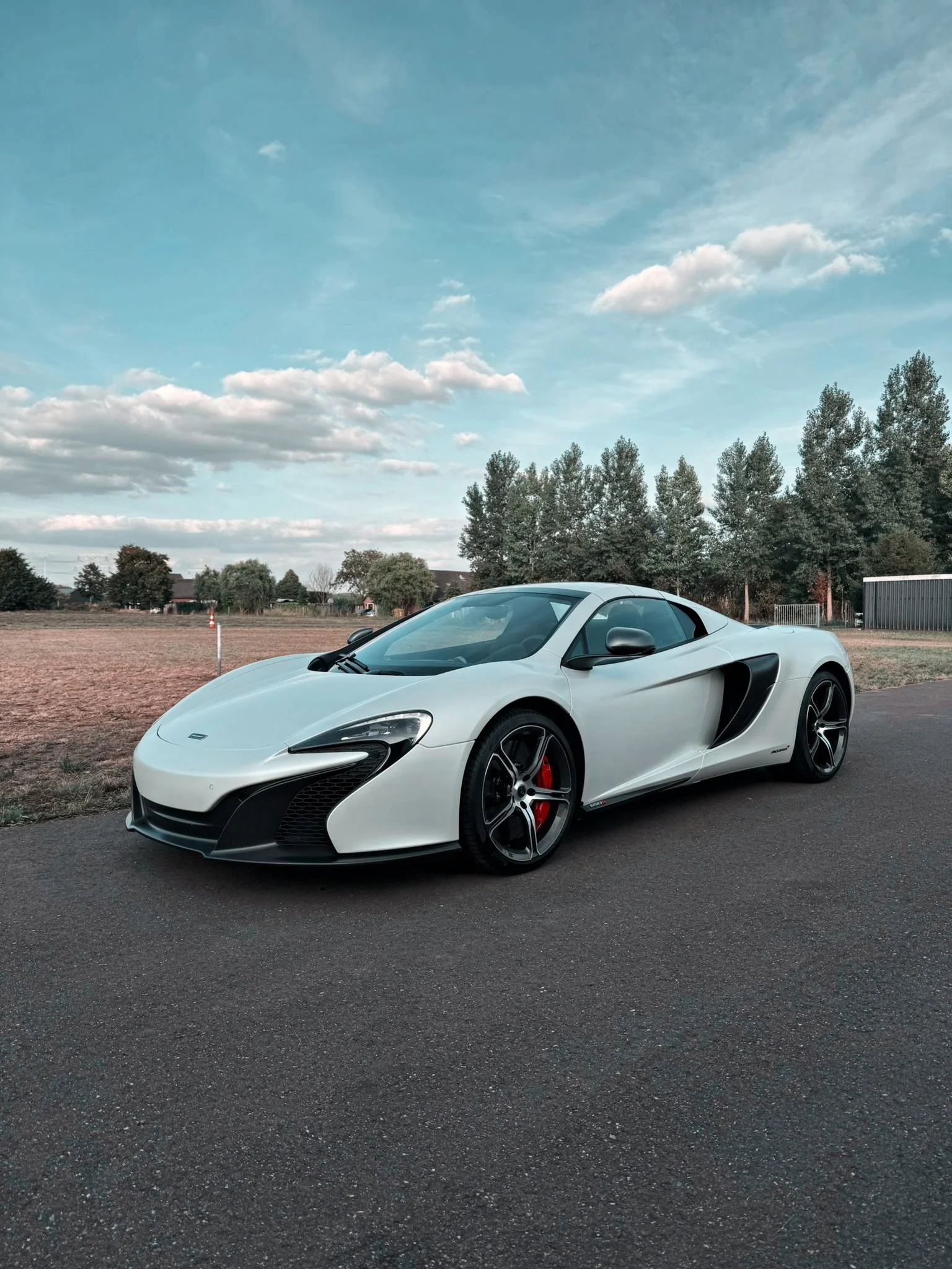 A white sports car with black accents parked on a paved road in a rural area with trees and a cloudy sky in the background.