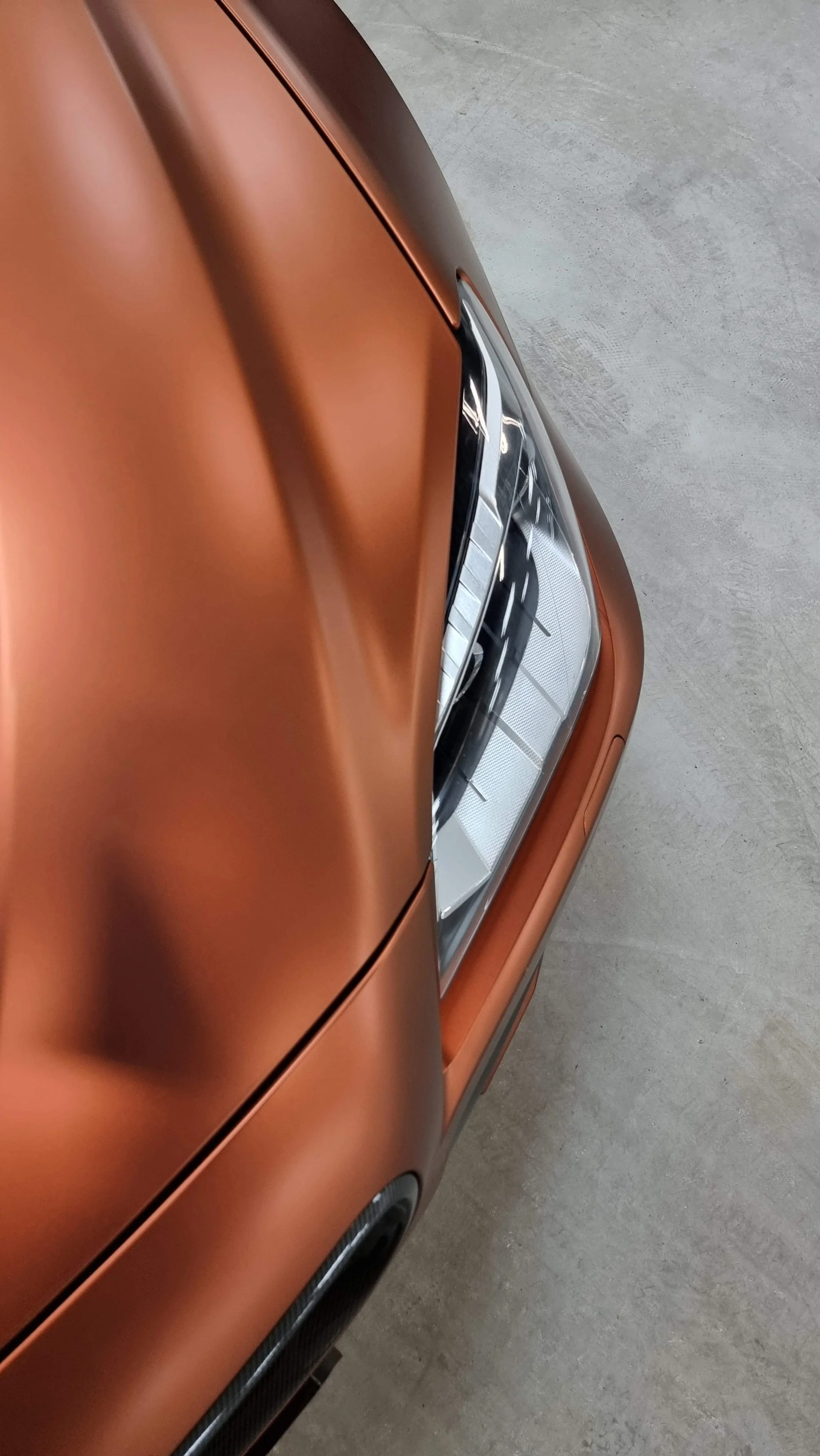 Close-up of the front end of a copper-colored sports car, showing part of the hood, headlight, and front grille.