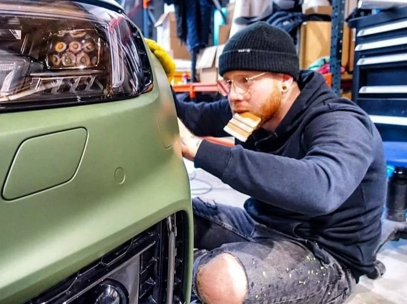 Man in black beanie and hoodie working on a green car in a garage.