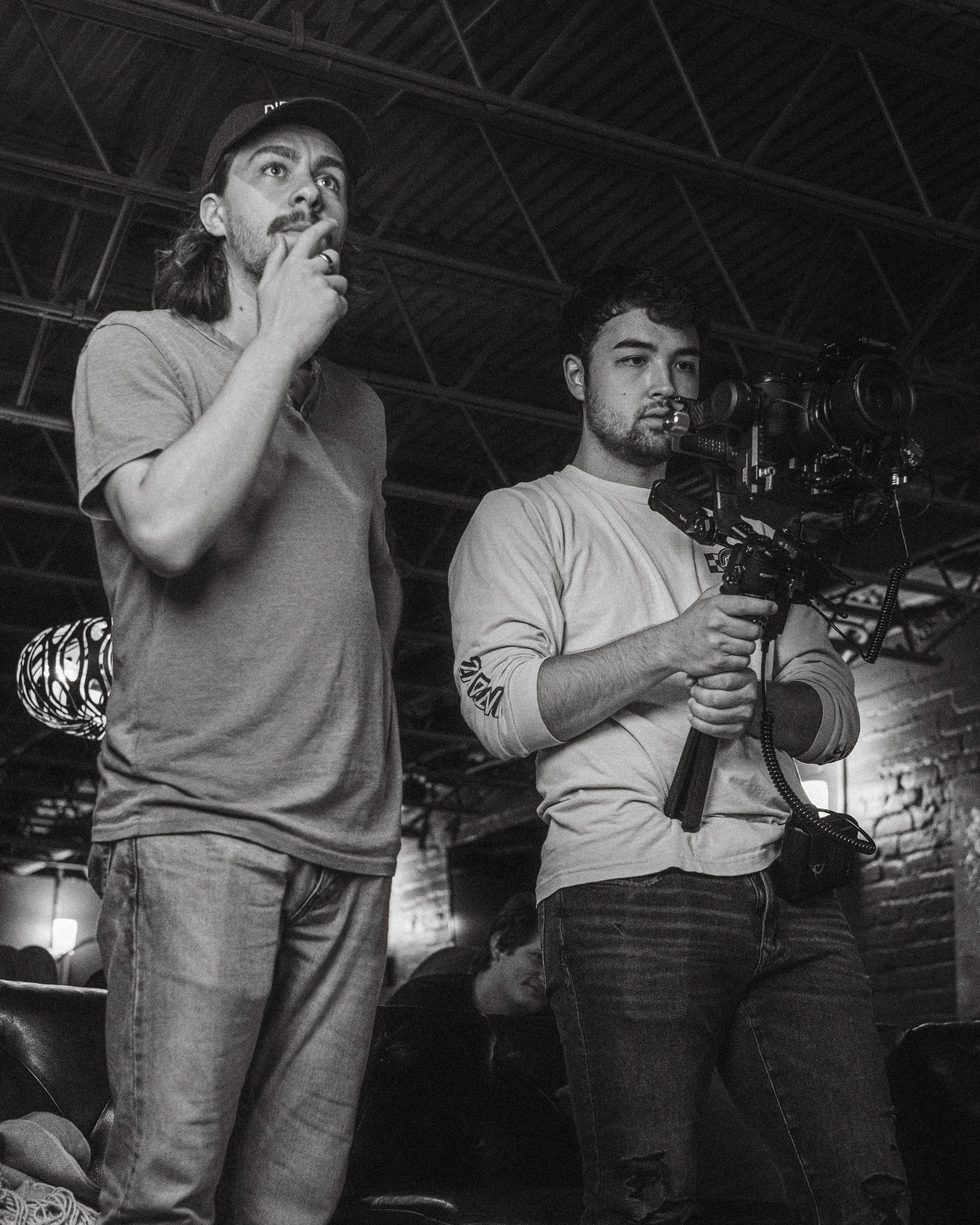 Two young men, one holding a professional video camera, stand side by side in a dimly lit indoor space with exposed brick walls and a metal ceiling. The man on the left wears a cap and casual t-shirt, looking thoughtful with his hand on his chin. The