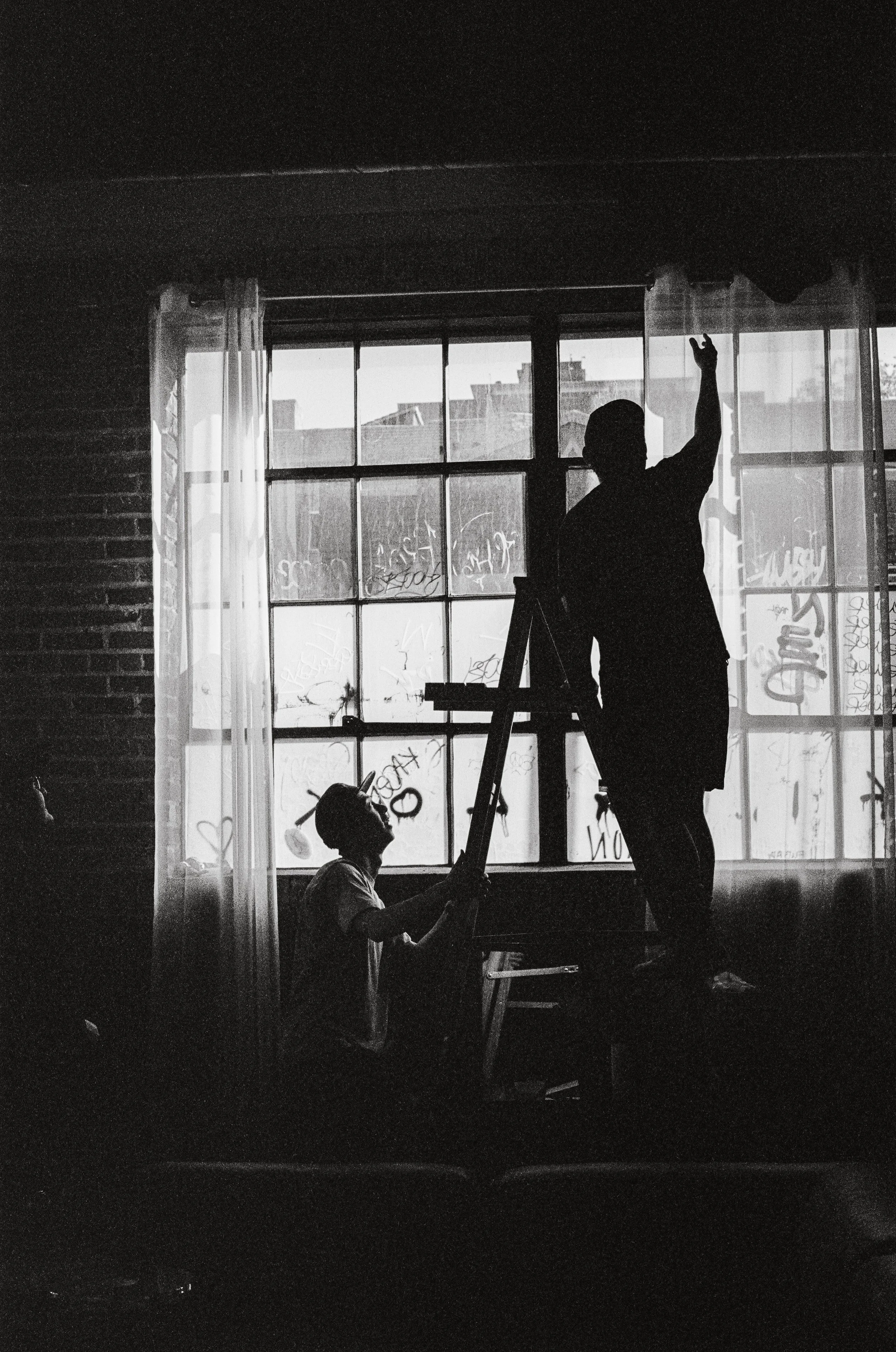 Silhouettes of two people, one standing on a ladder and the other sitting on a chair, near a window with graffiti on the glass. One person is reaching towards the window, and the scene is backlit with natural light.