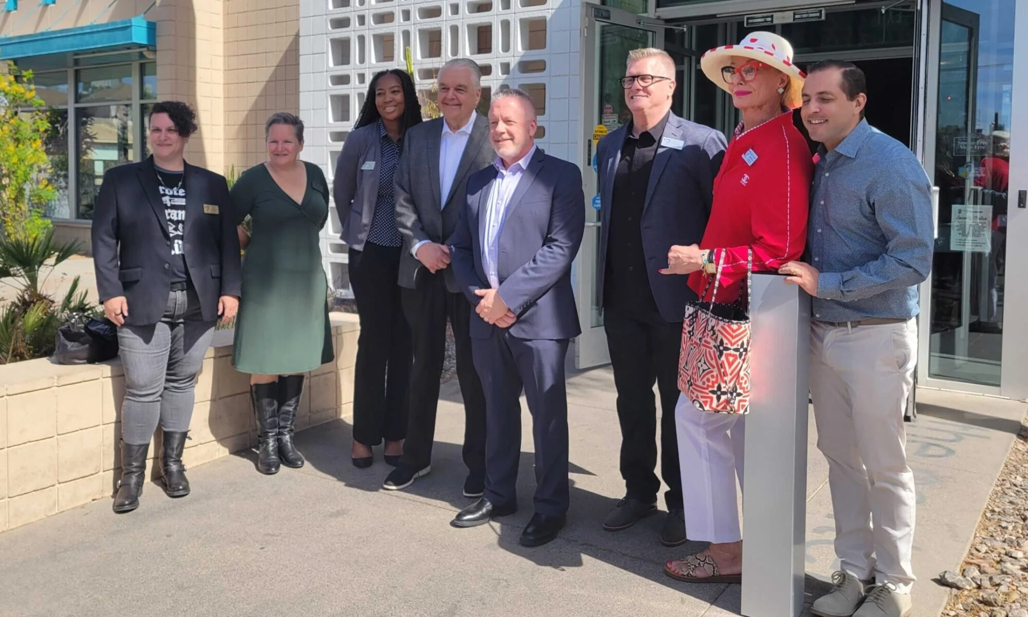 Group of eight diverse people standing outside in front of a building, posing for a photo during a sunny day.