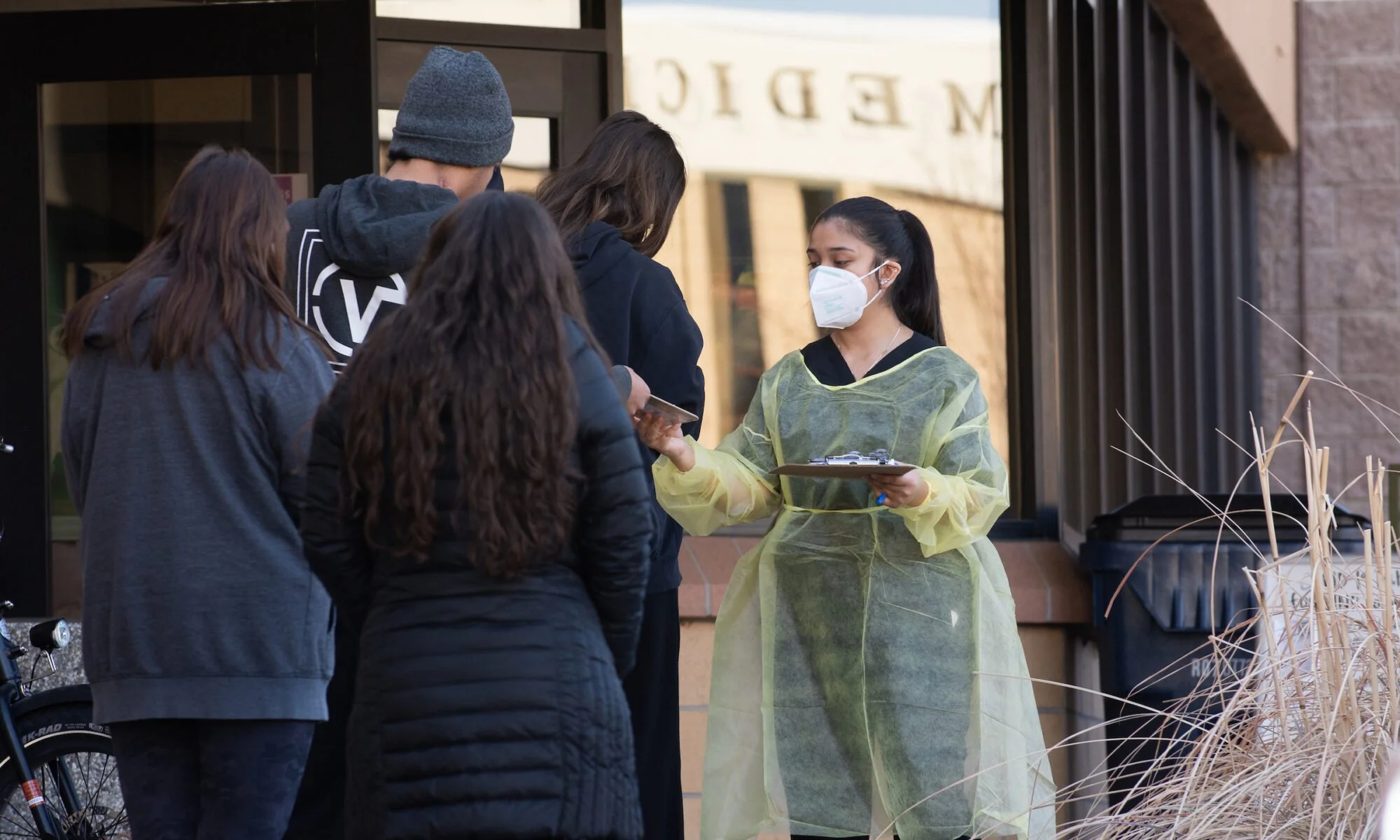 A healthcare worker in protective gown and face mask taking temperatures from a group of people outside a building.