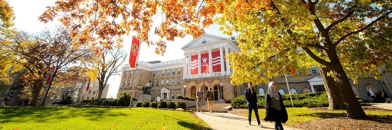Students walking on the campus lawn in front of a grand university building with fall foliage.