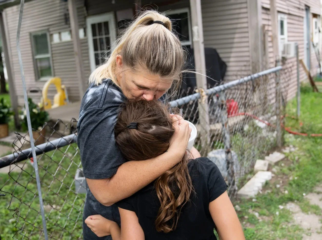 An emotional woman hugging a young girl in front of a neighborhood fence and house.