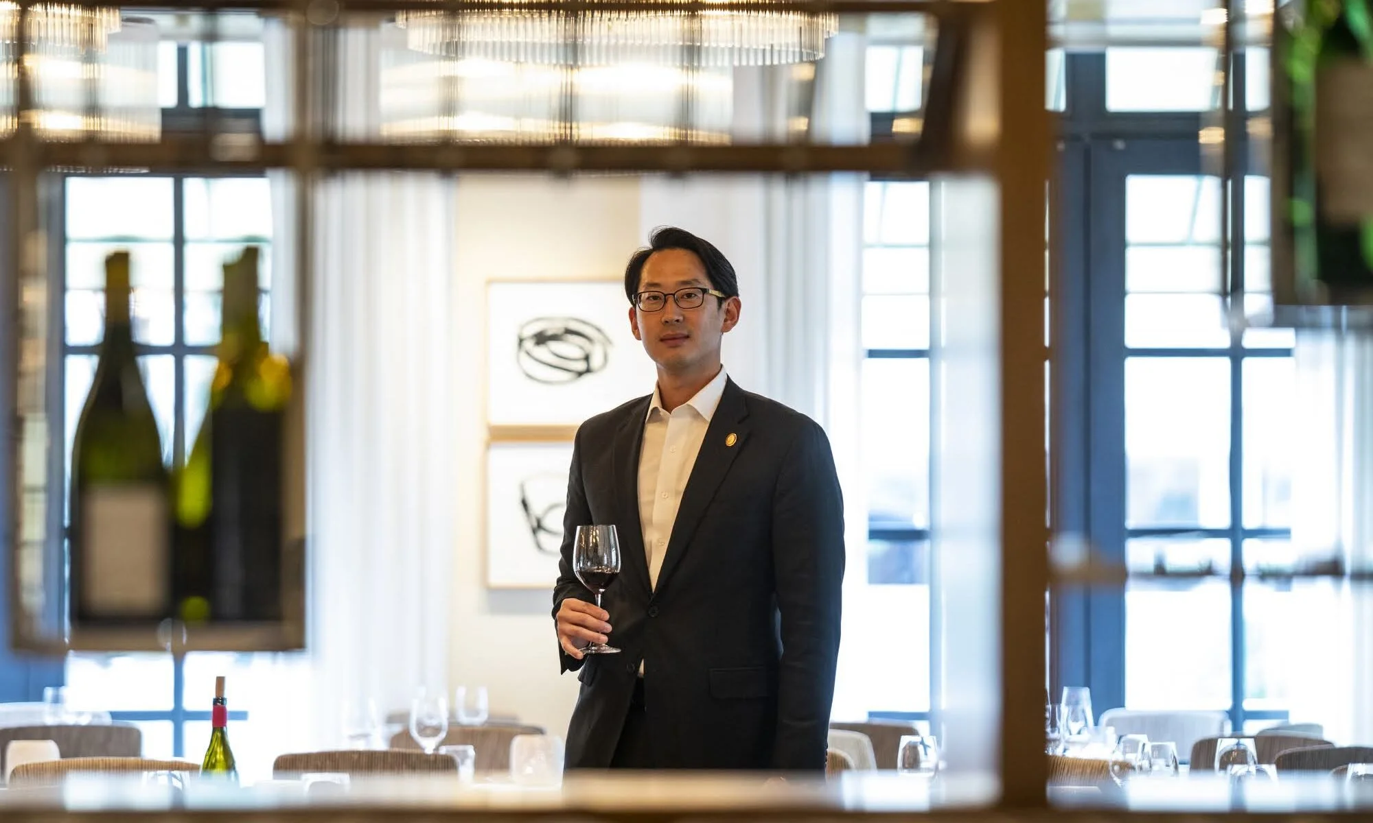 A man in a suit holding a glass of red wine in a restaurant or banquet hall with large windows and artwork on the wall.