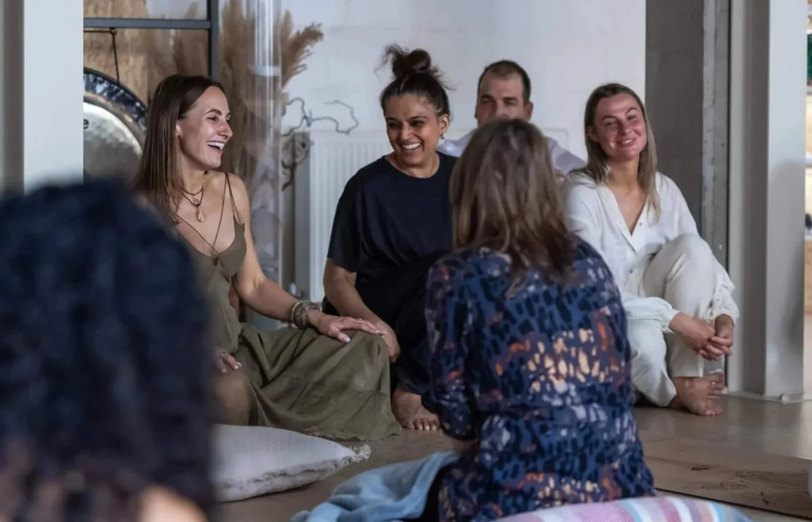 A group of diverse people sitting on the floor, smiling and engaging in conversation, in a cozy, modern indoor class at the Luv Yourself Studio, London.