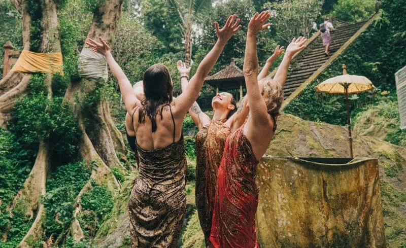 Three women reconnecting joyfully with arms raised in a lush outdoor setting, surrounded by trees, rocks, and a staircase in the background.