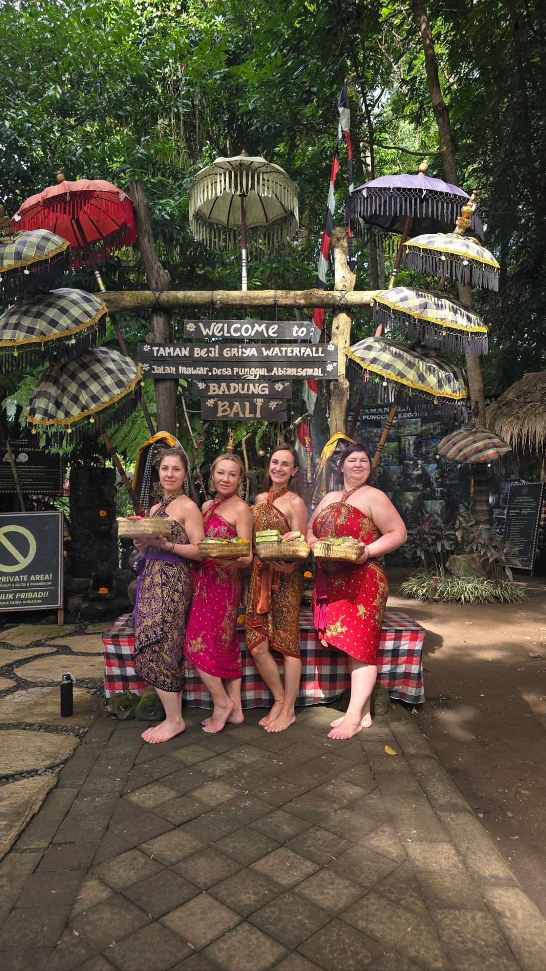 Four women in traditional Balinese attire holding baskets, standing in front of a sign and umbrellas at a cultural or tourist site in Bali, Indonesia.