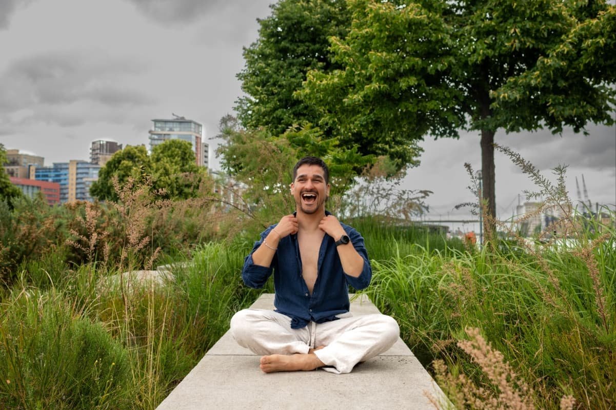 Our teacher is smiling and sitting outside in calm area of Royal Docks next to trees in modern surroundings