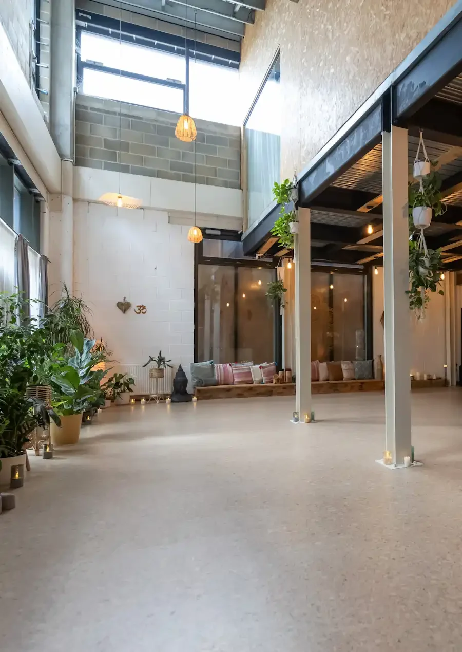 spacious yoga studio with natural light, wooden ceiling beams, hanging potted plants, and decorative items along the walls.
