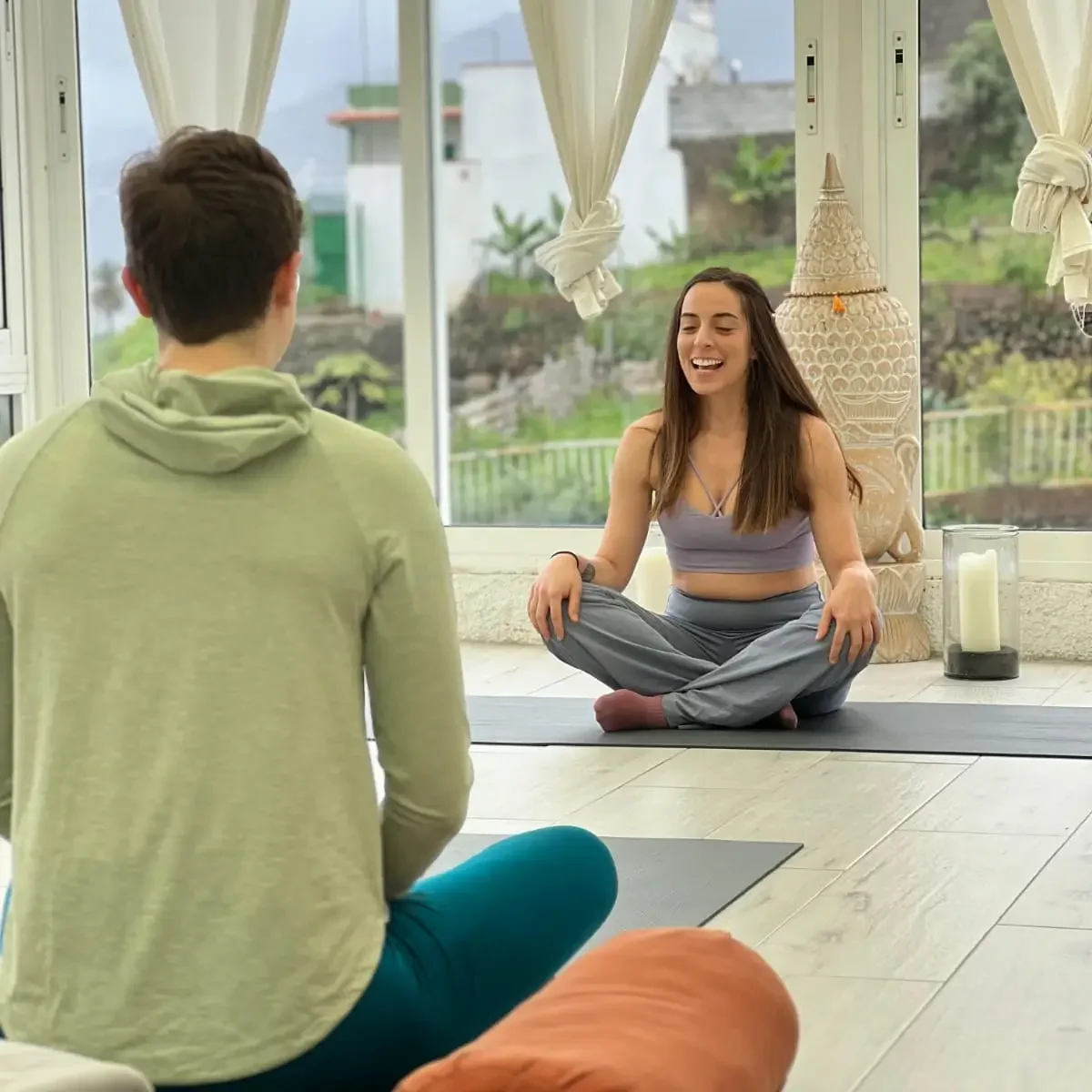 A smiling woman is sitting in a yoga pose in a class with another person.