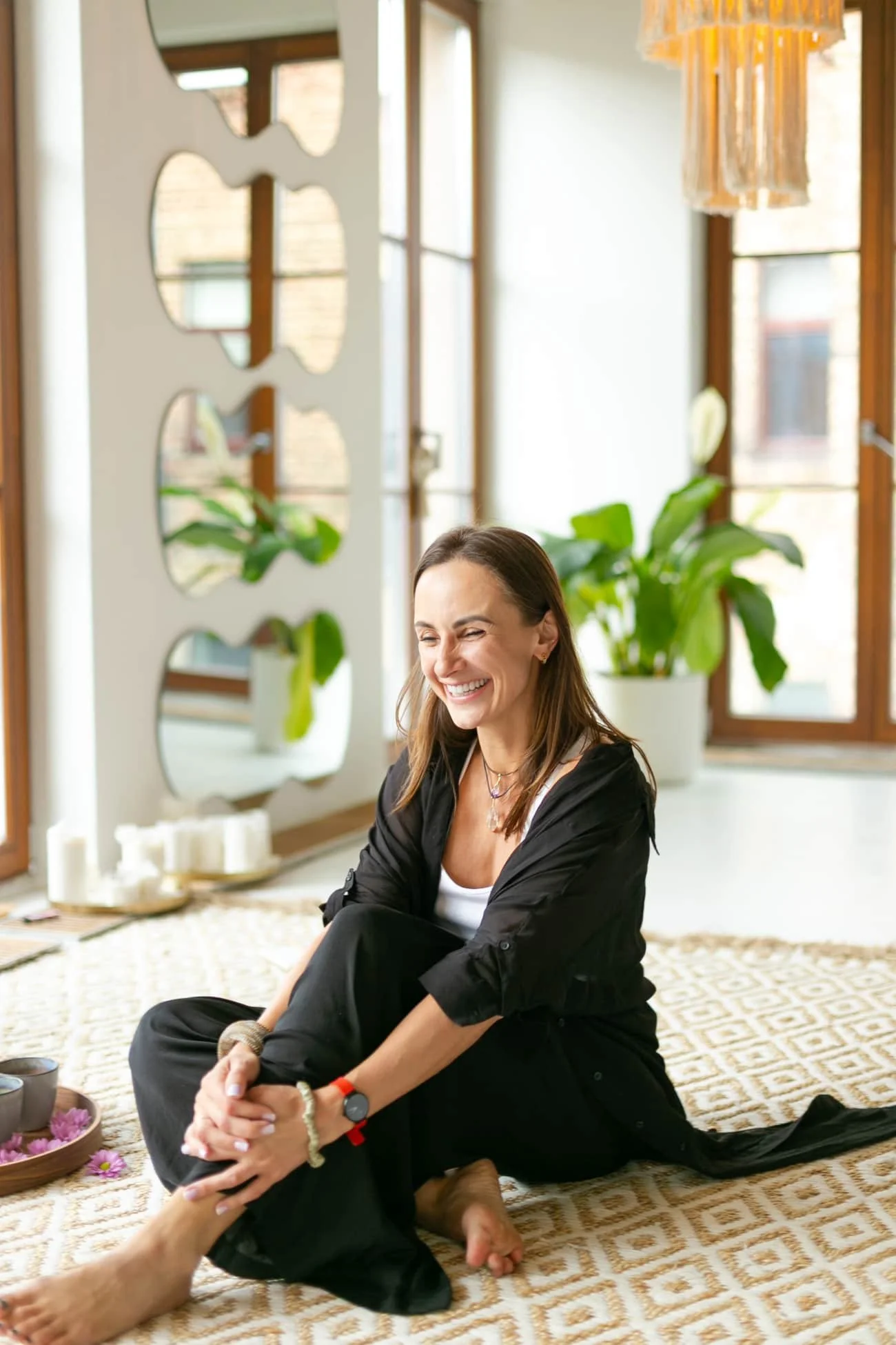 A woman sitting on a patterned beige and white rug in a bright room, smiling and laughing, surrounded by candles and potted plants after Life-Force Activation Facilitator training