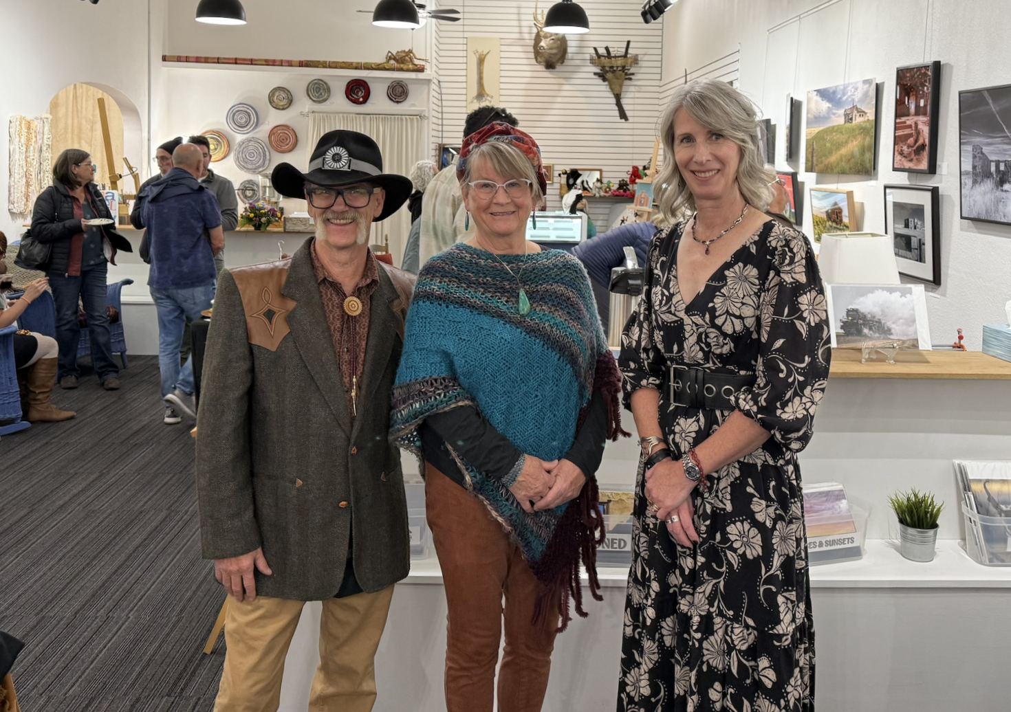 Three people standing together in an art gallery, smiling for the photo. The background shows other visitors, artwork, and decor on the walls.