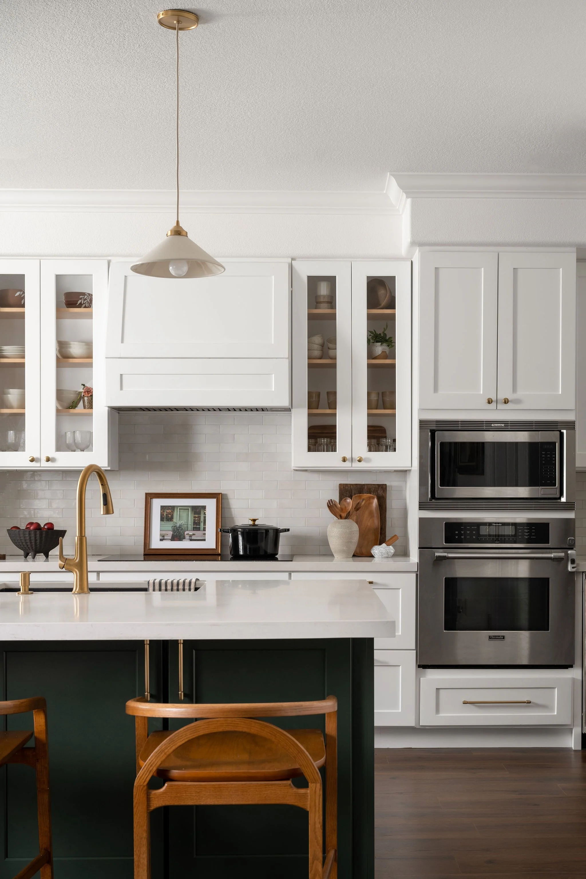 A modern kitchen with white cabinets, a white countertop, a brass faucet, and a kitchen island with a dark green base. Two wooden chairs are partially visible in the foreground. There's a metallic microwave oven and a wall with framed artwork behind 