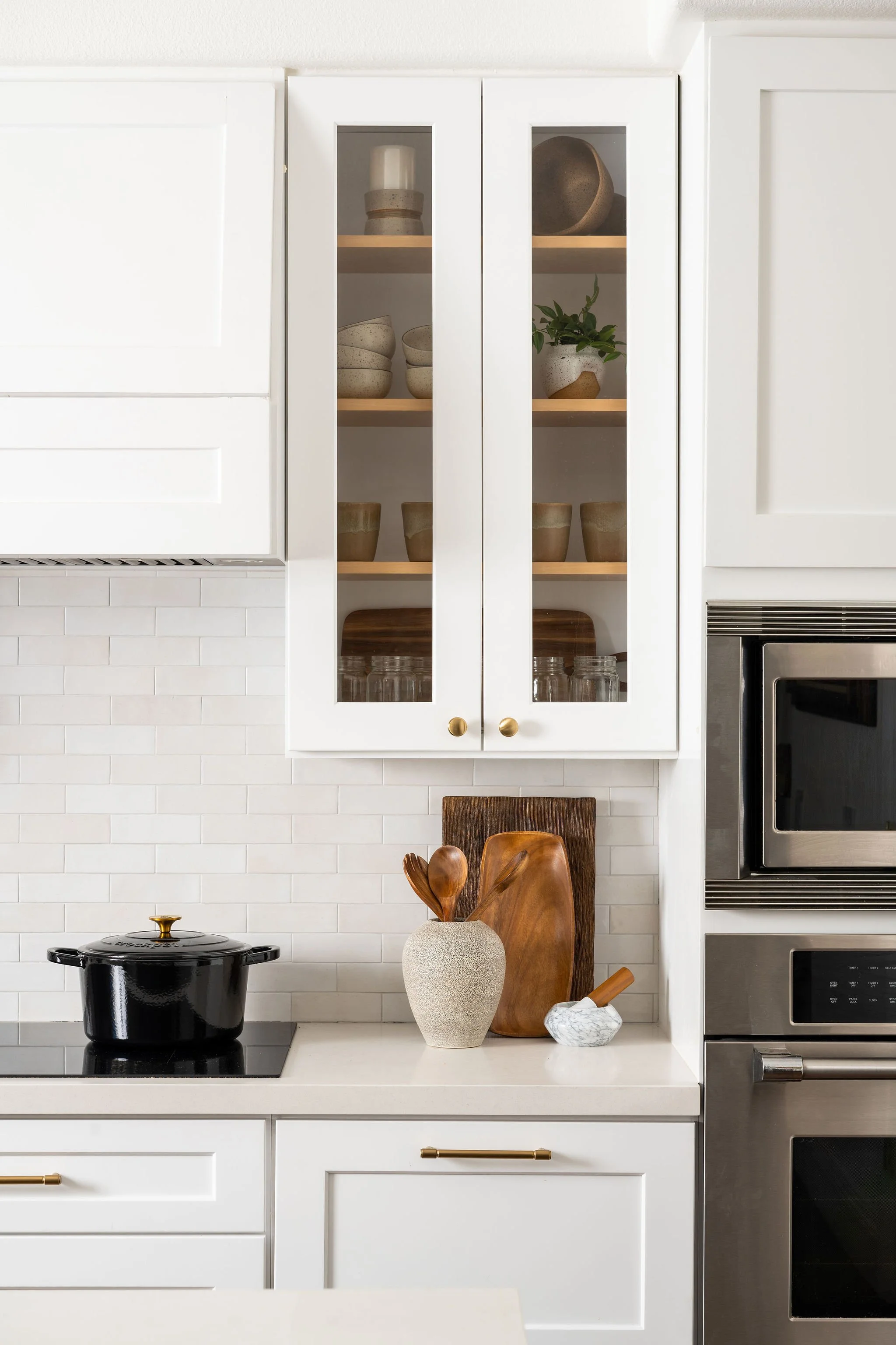 White kitchen cabinet with glass doors displaying ceramic bowls, a potted plant, and other kitchenware. On the counter below, there are decorative wooden utensils in a white vase, a black pot, and a small marble dish. The background features a white 