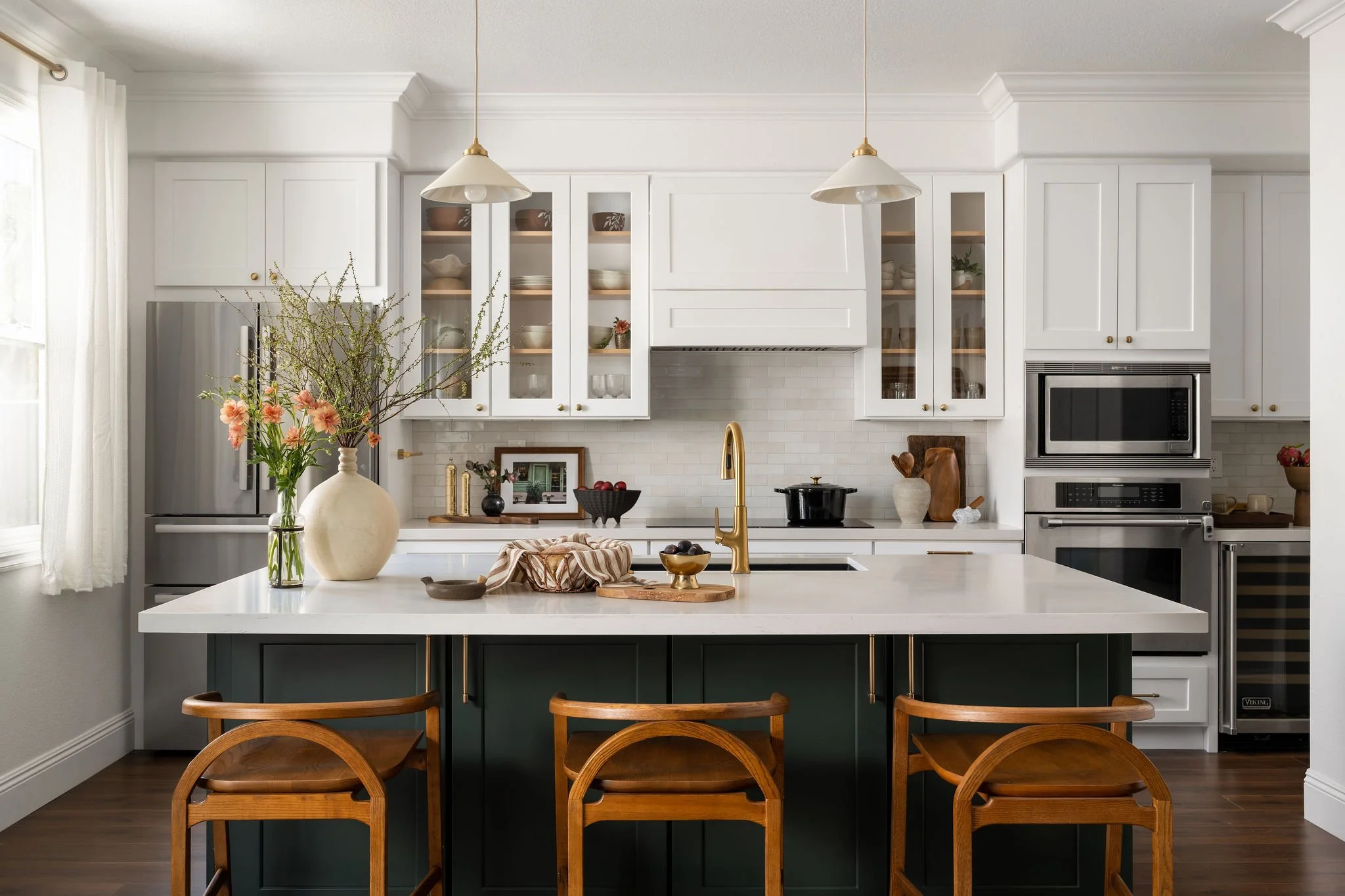 Modern white kitchen with open shelving, stainless steel appliances, and a large island with a green base and wooden chairs. Decor includes flowers in a white vase and kitchen utensils.