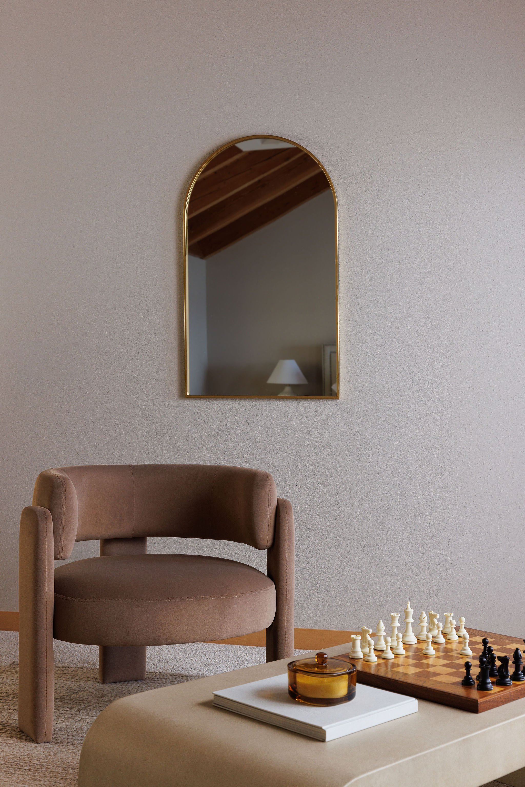 Interior of a living room with a plush beige armchair, a low coffee table with a chess set, a candle, and a stack of books, a gold-framed mirror on the wall reflecting a wooden ceiling and lamp.