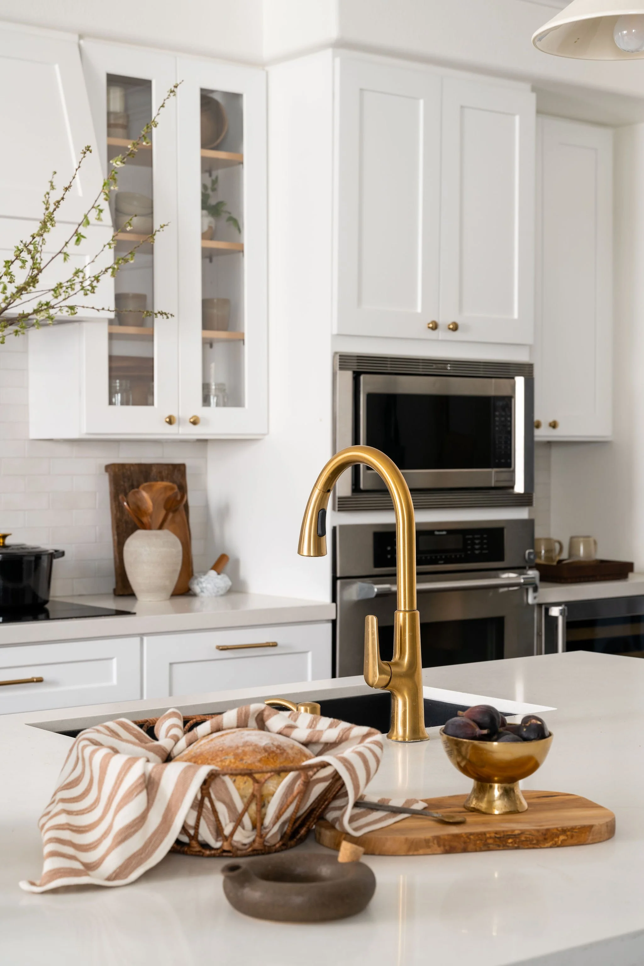 A modern kitchen with white cabinets, a gold faucet, a microwave oven, a bowl of fruit, a bread basket with bread and a striped dish towel on a kitchen island.