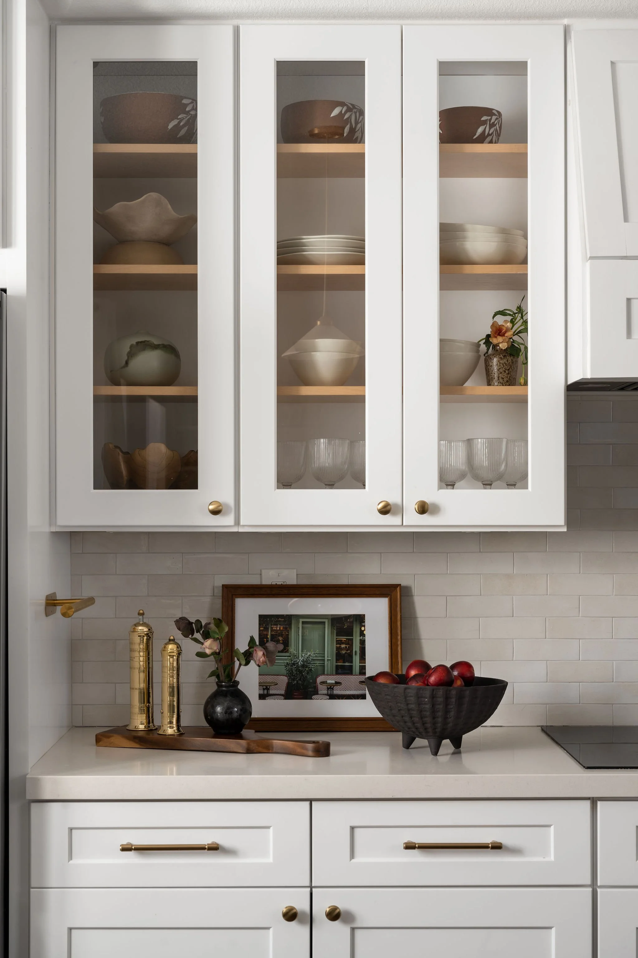 White kitchen cabinet with glass doors displaying ceramic bowls and plates, decorated with flowers and art pieces on the countertop below.
