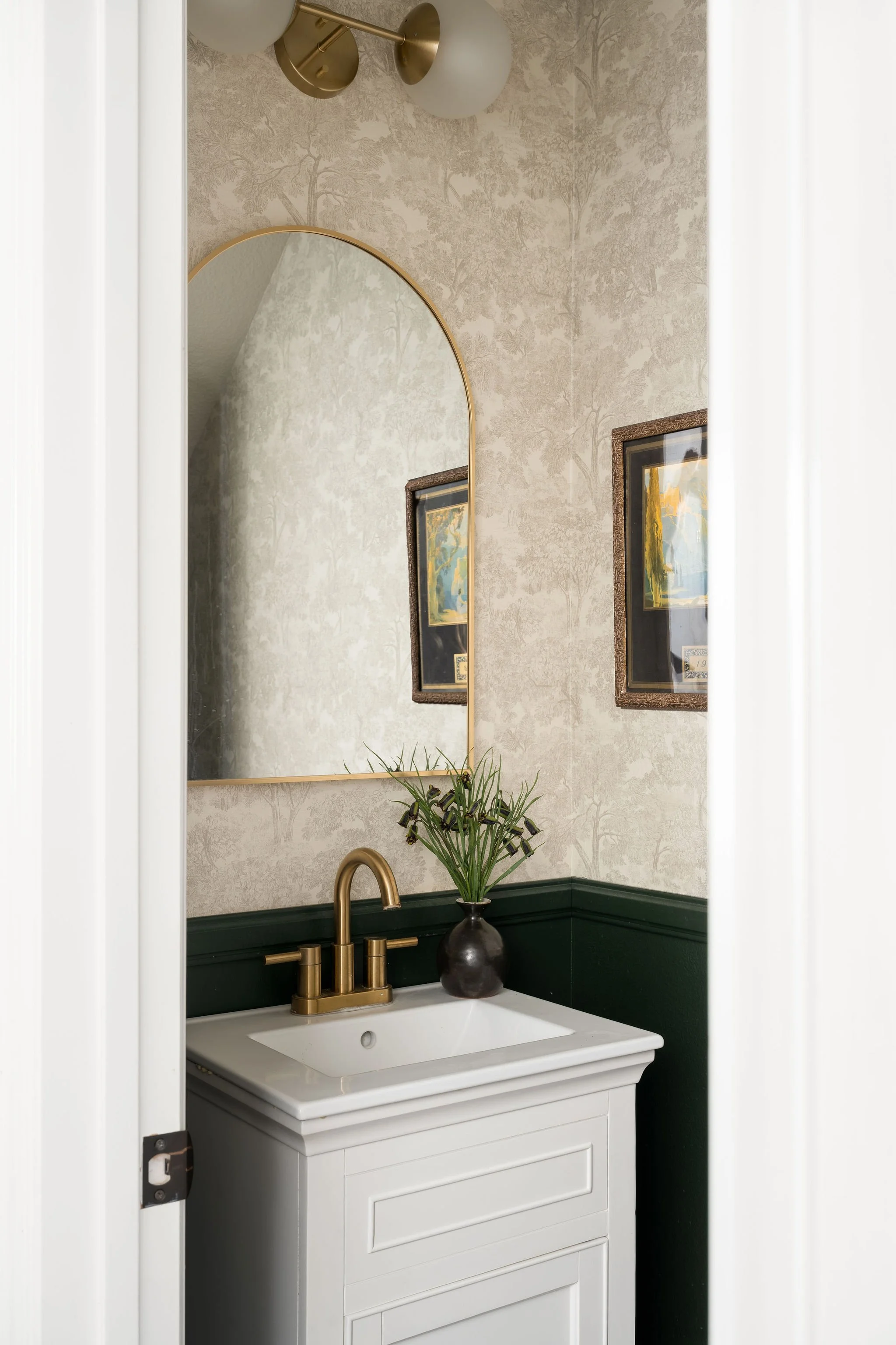 A bathroom vanity with a white sink and cabinet, a gold faucet, a dark green backsplash, a dark vase with greenery, a gold-framed mirror, wallpaper with tree patterns, and framed artwork on the wall.