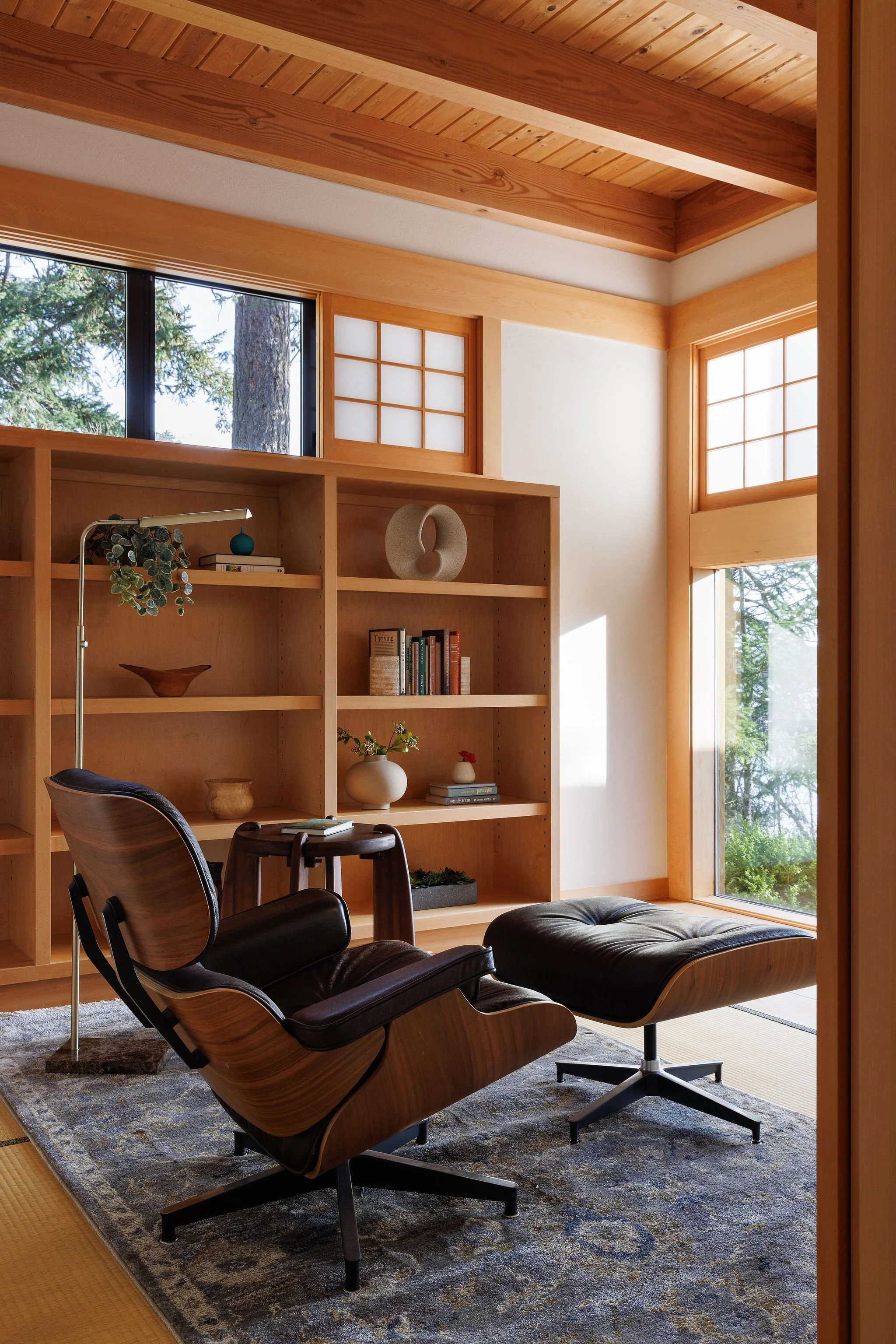 Interior of a room with wooden ceiling and large windows, featuring a black leather lounge chair with matching ottoman on a decorative rug, and a wooden bookshelf with decorative items and books.