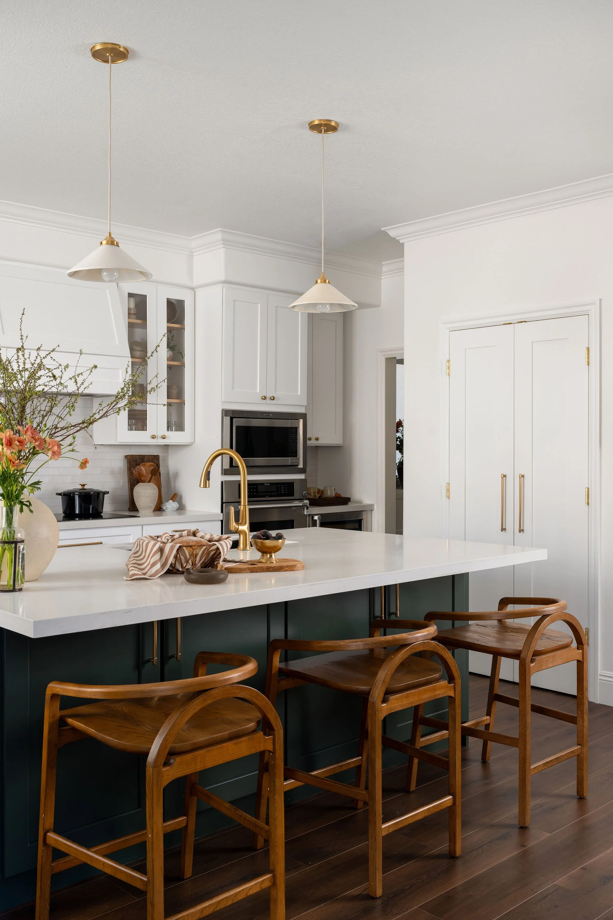 A modern kitchen with a large white island counter, wooden chairs, white cabinetry, gold fixtures, and hanging pendant lights.