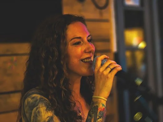 A woman with curly hair singing into a microphone in a dimly lit setting.