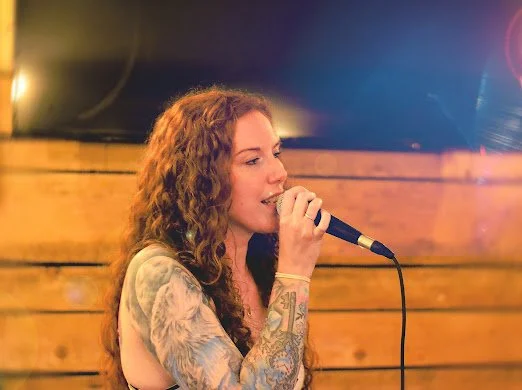 A woman with curly red hair, tattoos on her arm, singing into a microphone in front of a wooden wall background.
