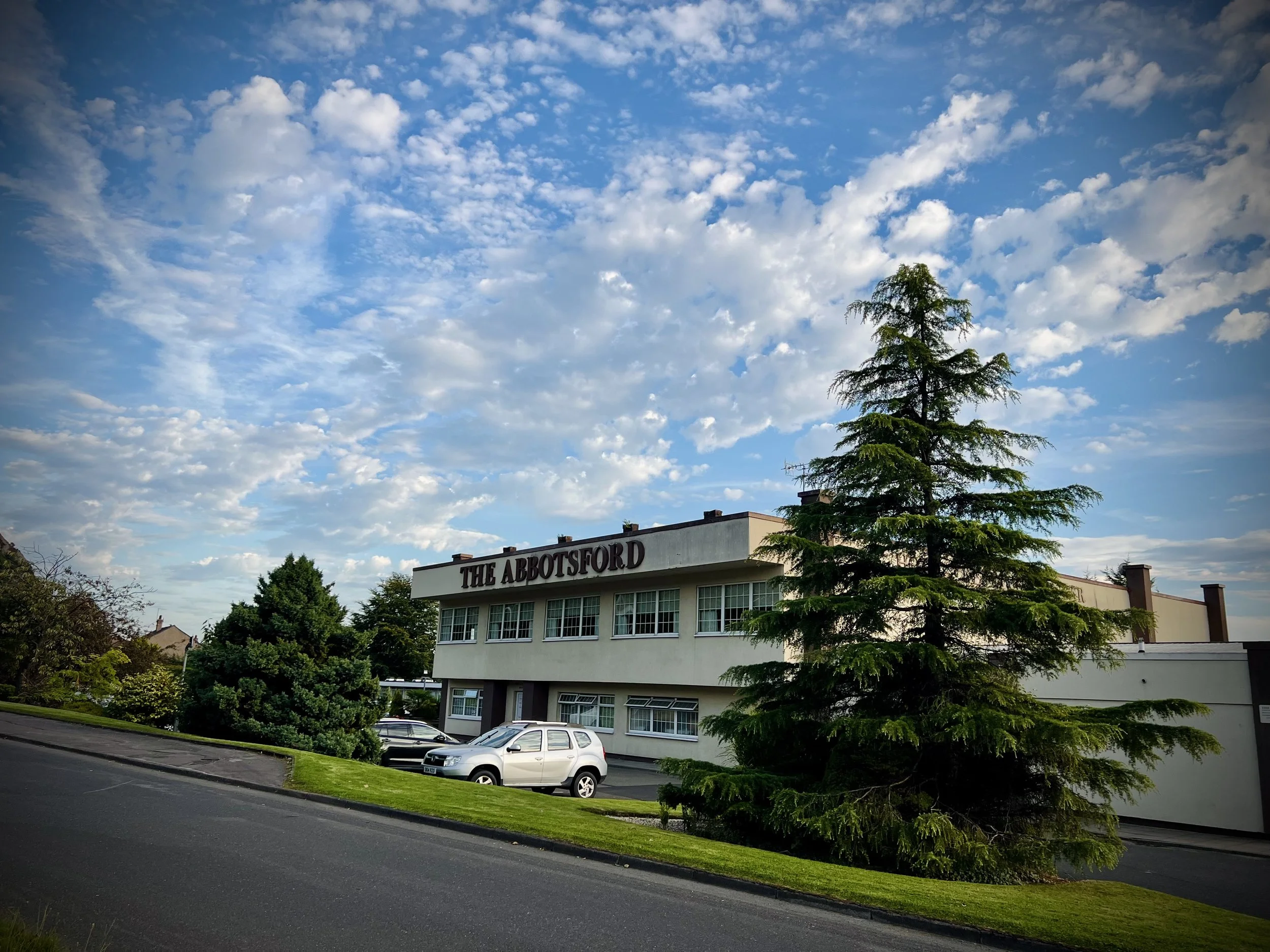 Exterior photo of the Abbotsford Hotel, a rectangular light stone coloured building with the words ABBOTSFORD spelled out at the top of the building. In the foreground you can see some cars and a tree. In the background is a bright blue sky and cloud
