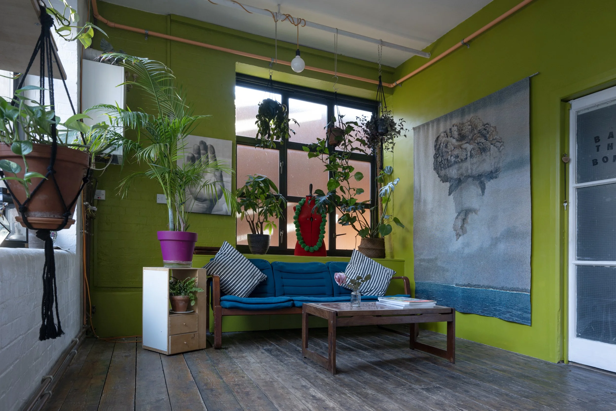 Living room with green walls, a blue sofa with black-and-white pillows, a wooden coffee table, various potted plants, colorful wall art, and a large window.