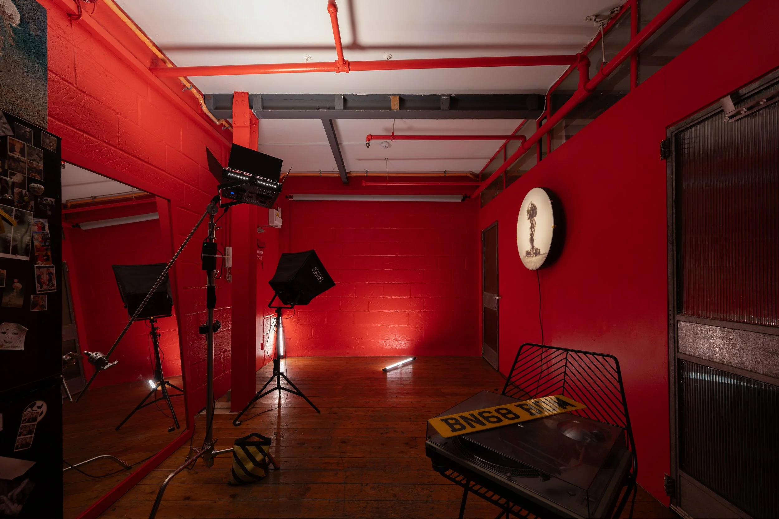 A room with bright red painted brick walls, wooden floor, and various studio equipment including lights, a mirror, a turntable, and a yellow and black striped license plate.