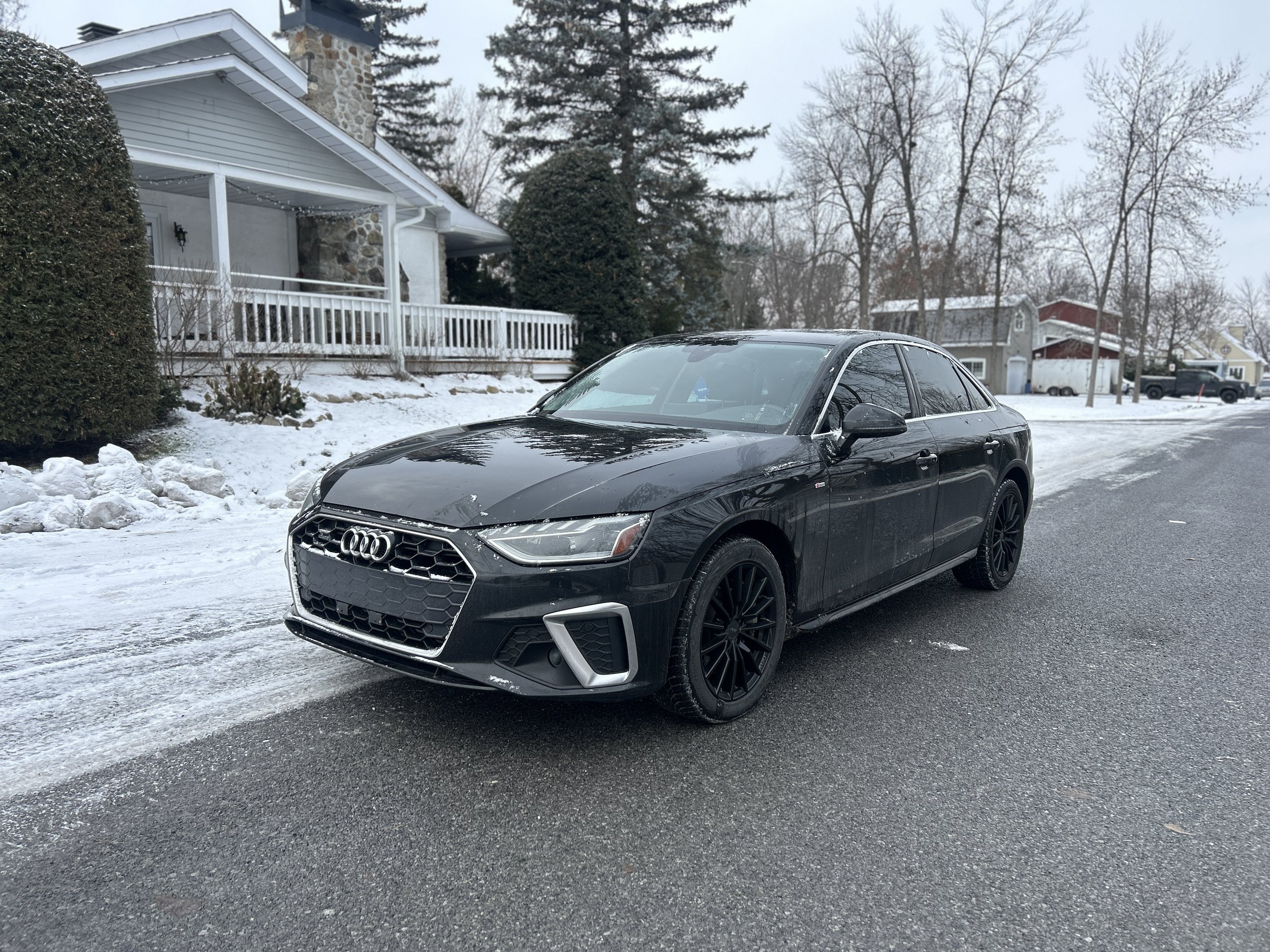 A black Audi sedan parked on a snowy street with snow around the tires, in a residential neighborhood with houses, trees, and a cloudy sky.