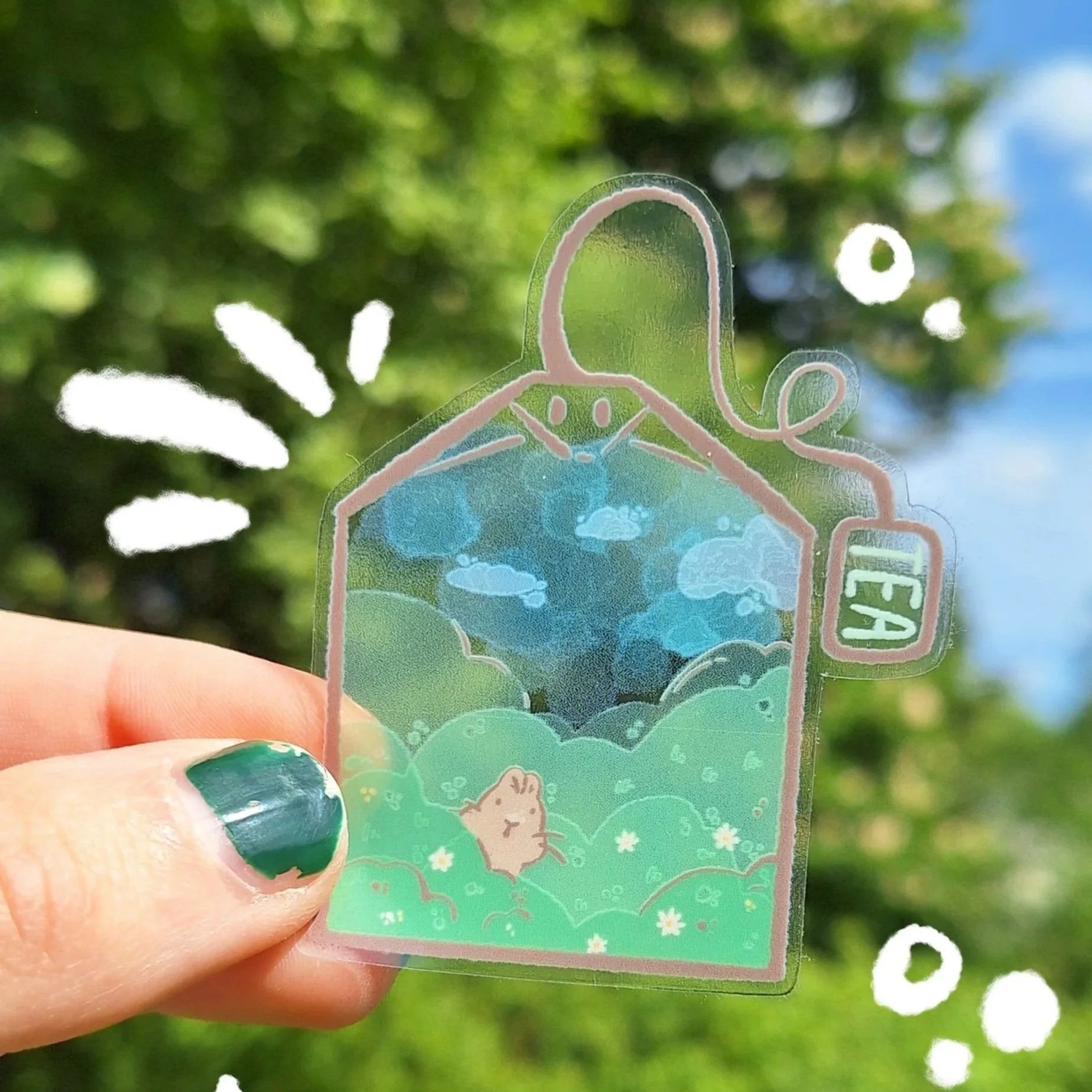 A transparent  tea bag shaped sticker with a landscape scene and bunnies, held by a person's hand with green nail polish, against a background of trees and blue sky with clouds.