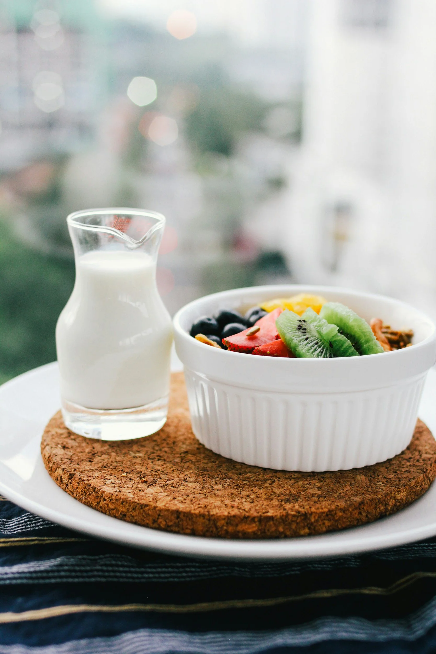 A bowl of mixed fruit and granola, a small pitcher of milk, and a cork coaster on a white plate, with a blurred cityscape background.